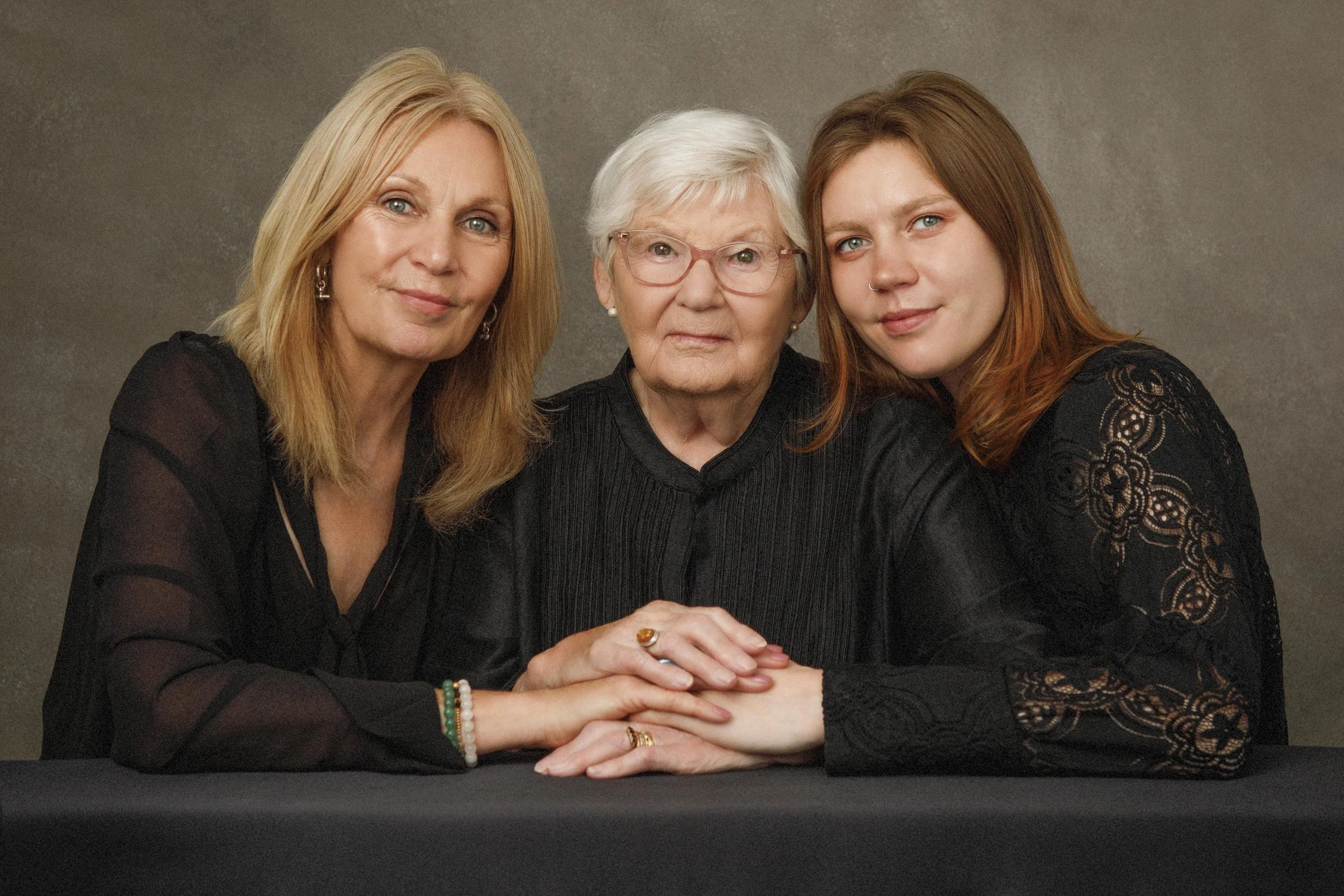 Three women sitting at a table, holding hands, looking at the camera, with neutral expressions, against a plain background.
