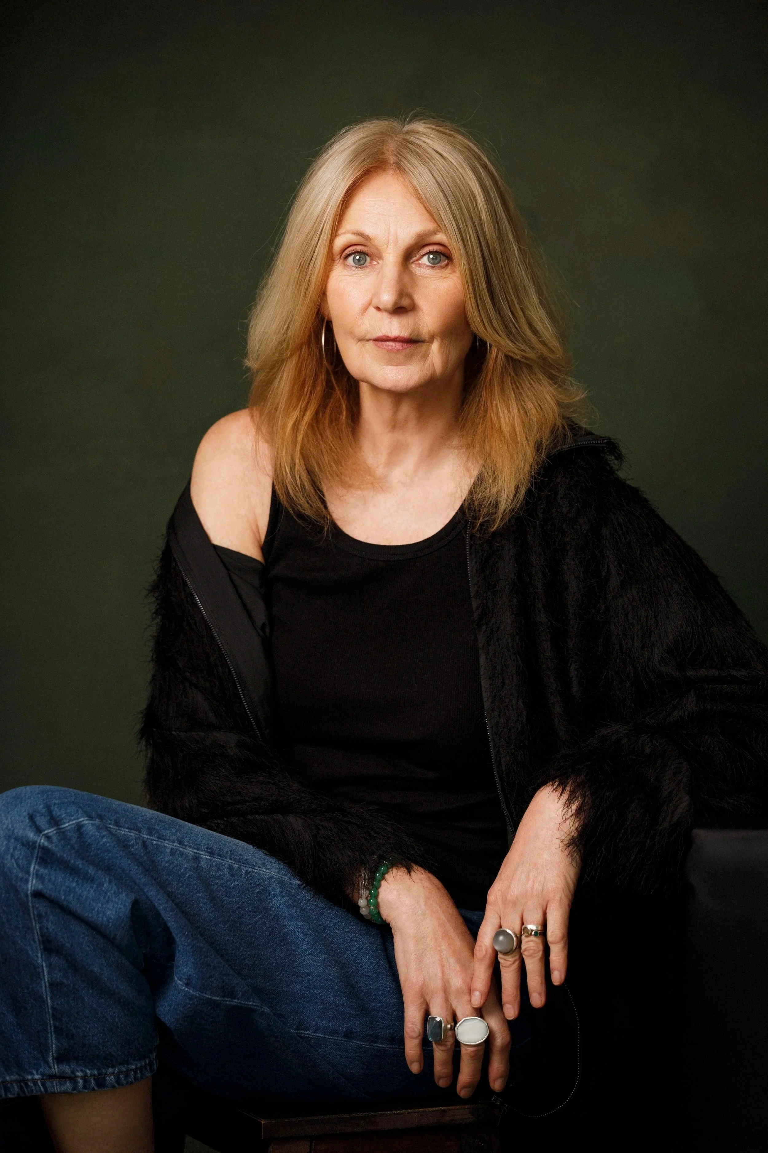 A woman with shoulder-length light brown hair and blue eyes, wearing a black top and black furry jacket, sitting against a dark green background. She has a calm expression and is accessorized with rings, a bracelet, and hoop earrings.