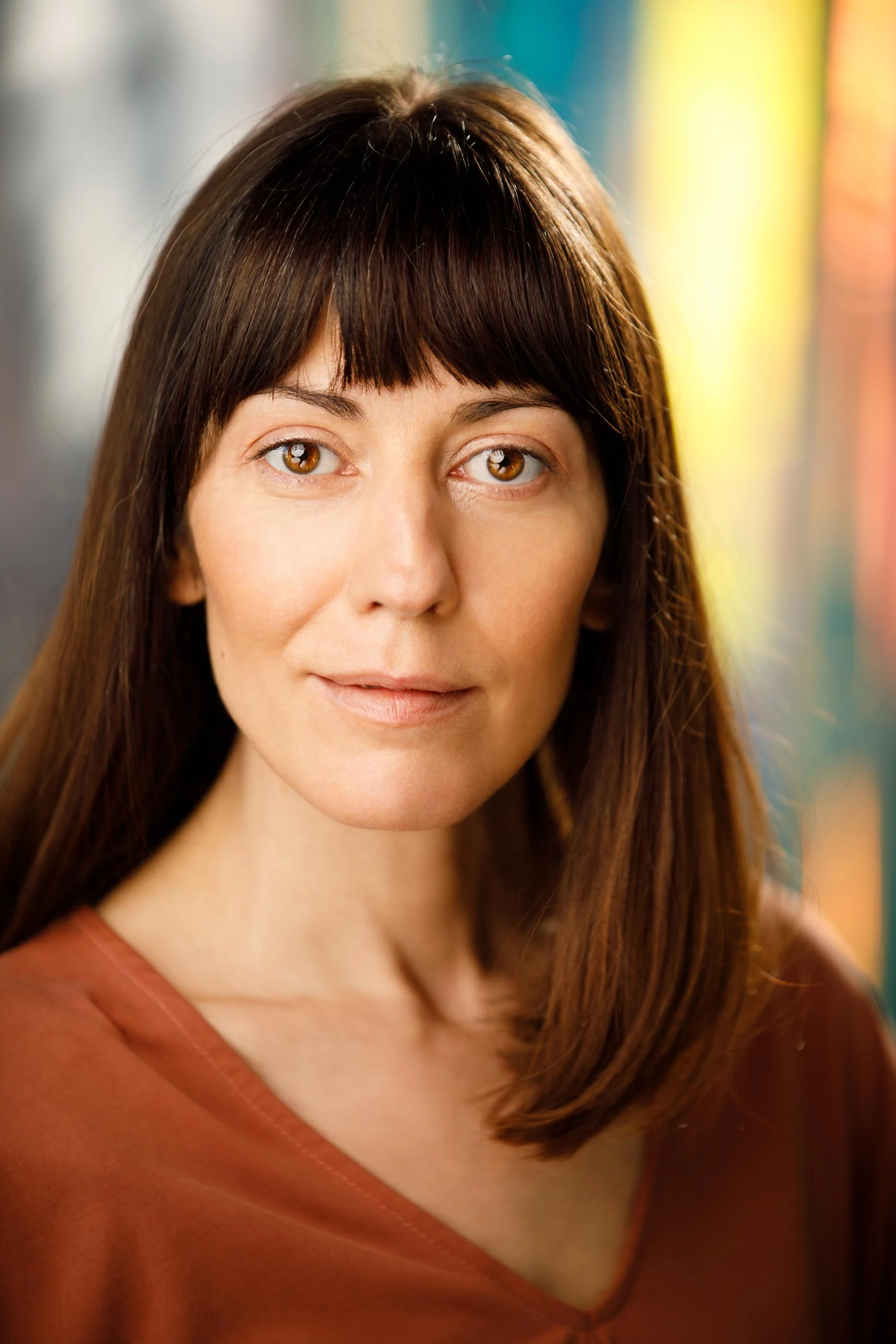 A close-crop portrait of a woman with a dark fringe and long straight hair, wearing a rust-coloured top against a softly blurred colourful backdrop. Minimal makeup, natural skin, direct and unflinching gaze. No artifice, no over-styling — just precis