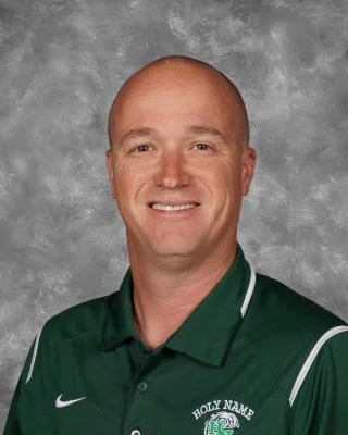 A smiling man with a shaved head wearing a green team polo shirt with white accents, logo, and text reading 'Holy Name.'