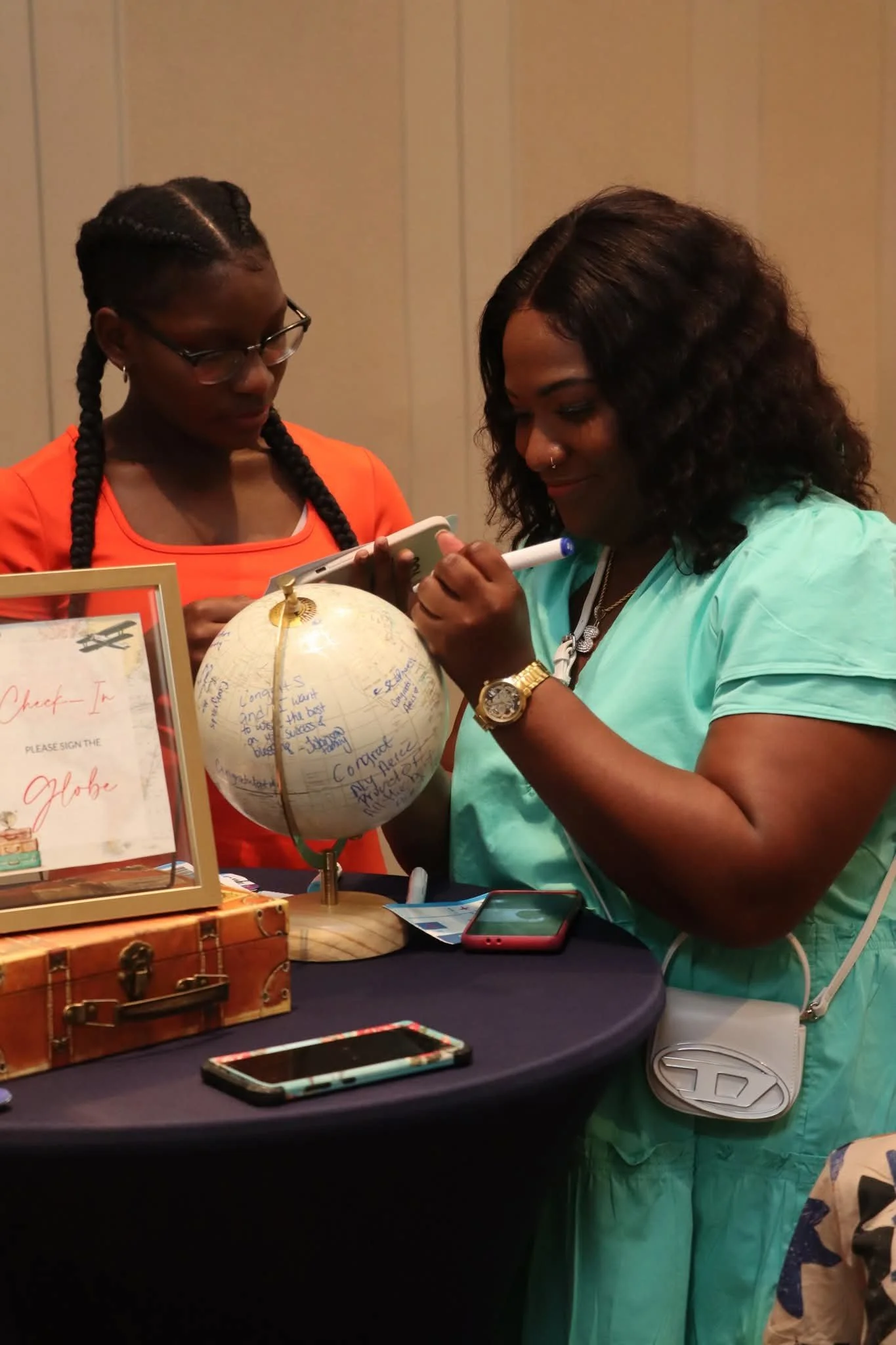 Two women are signing a globe at an event table. One woman is wearing glasses and an orange top, while the other is in a mint green shirt with a white purse. The table has a sign, phones, and a decorative box.