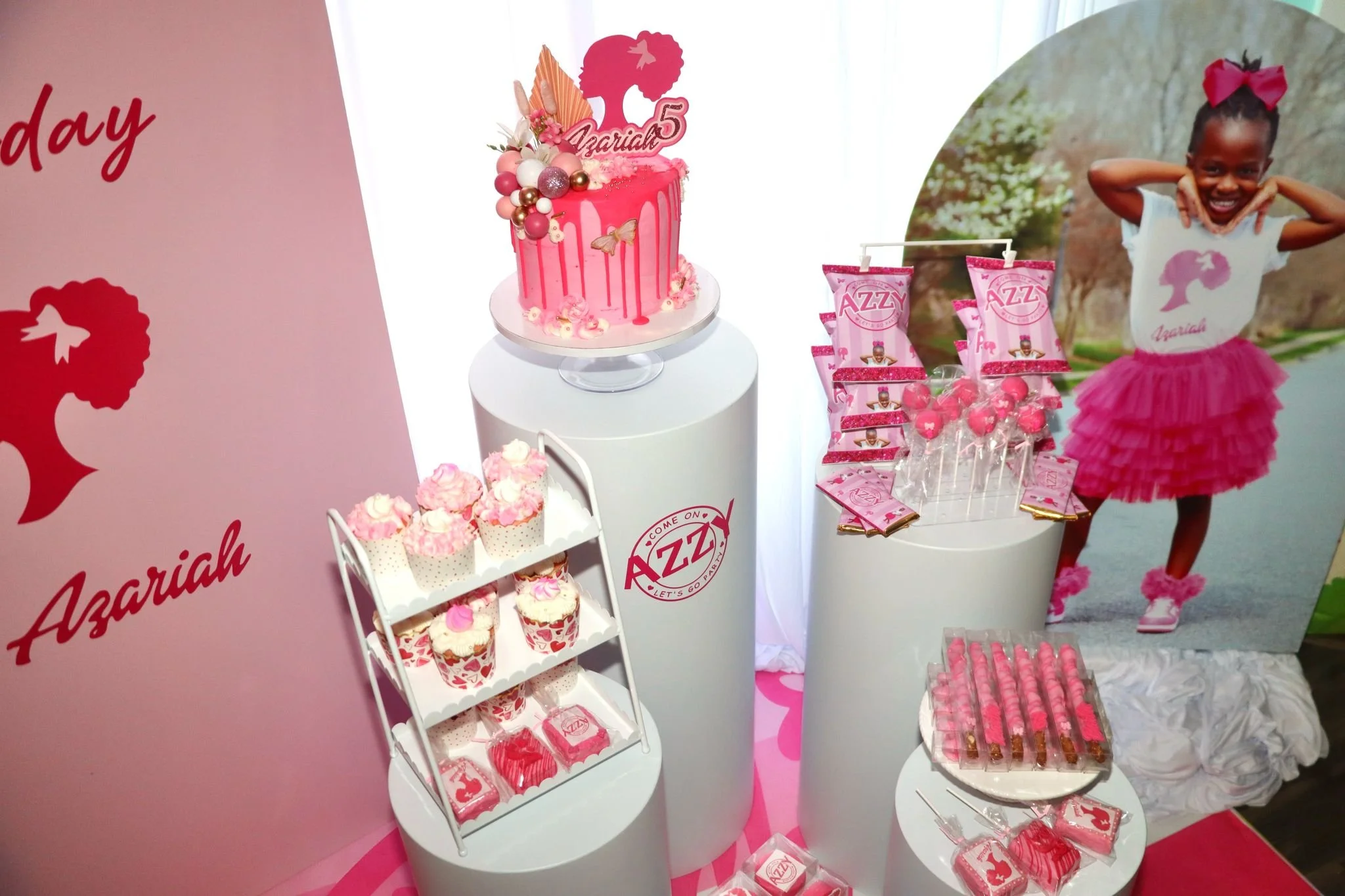 A pink-themed birthday party setup with a decorated cake, cupcakes, and treats, featuring a large photo of a smiling girl in a pink tutu and a shirt with a silhouette of a girl and the name Azariah. The decorations include pink and white balloons, an