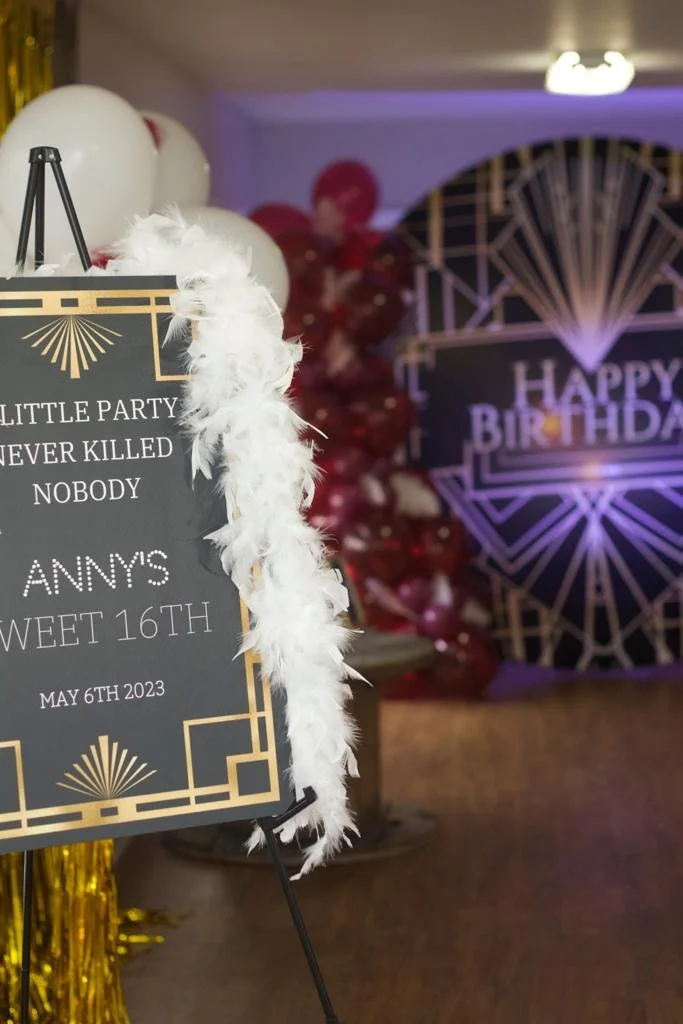 Party sign with gold accents and white feather trim; balloons and a decorated space with a "Happy Birthday" sign in the background for a birthday celebration on May 6th, 2023.