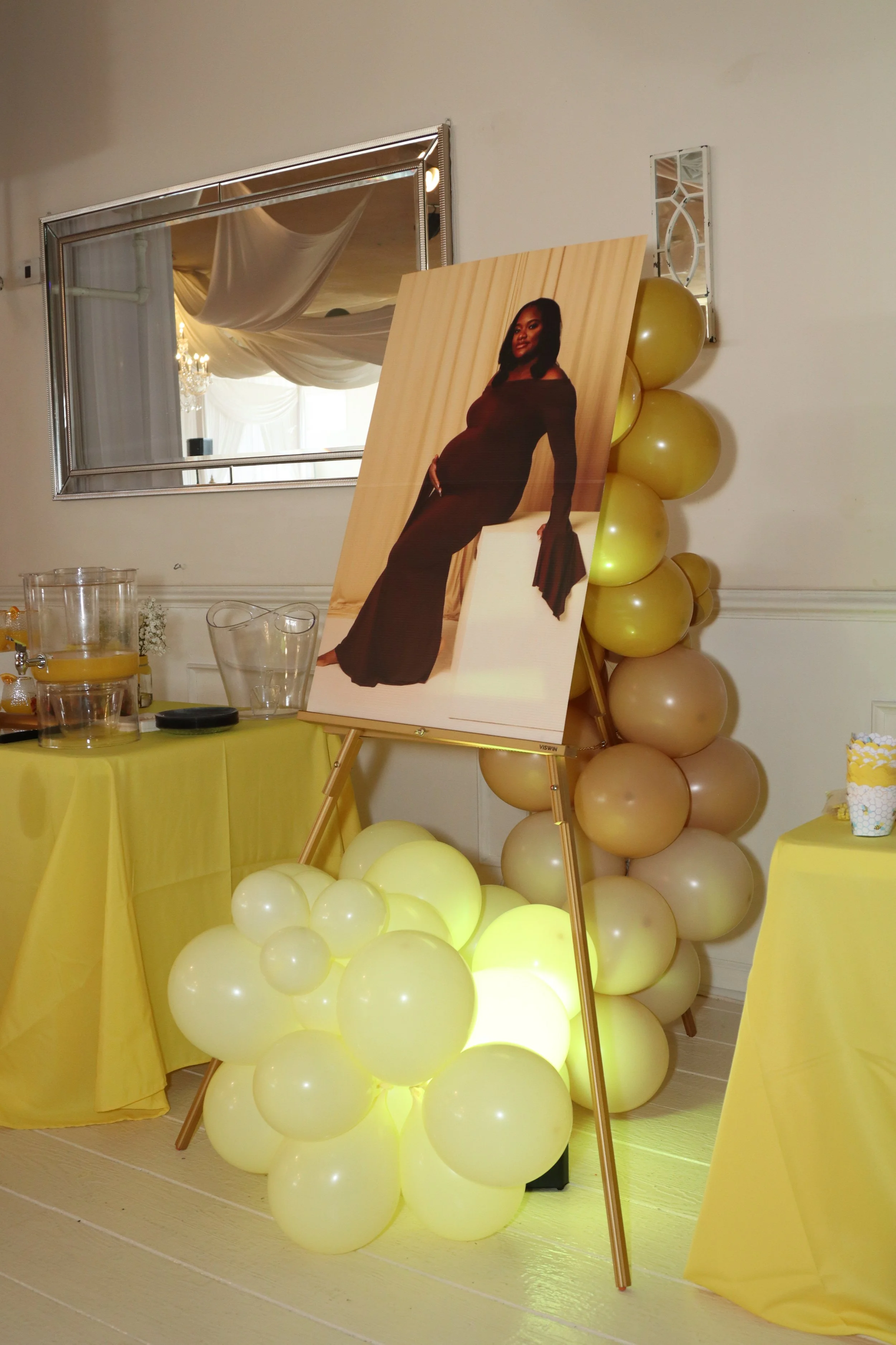 Decorative display with a large portrait of a pregnant woman in a dark dress, balloons, and yellow tablecloths at an indoor celebration.