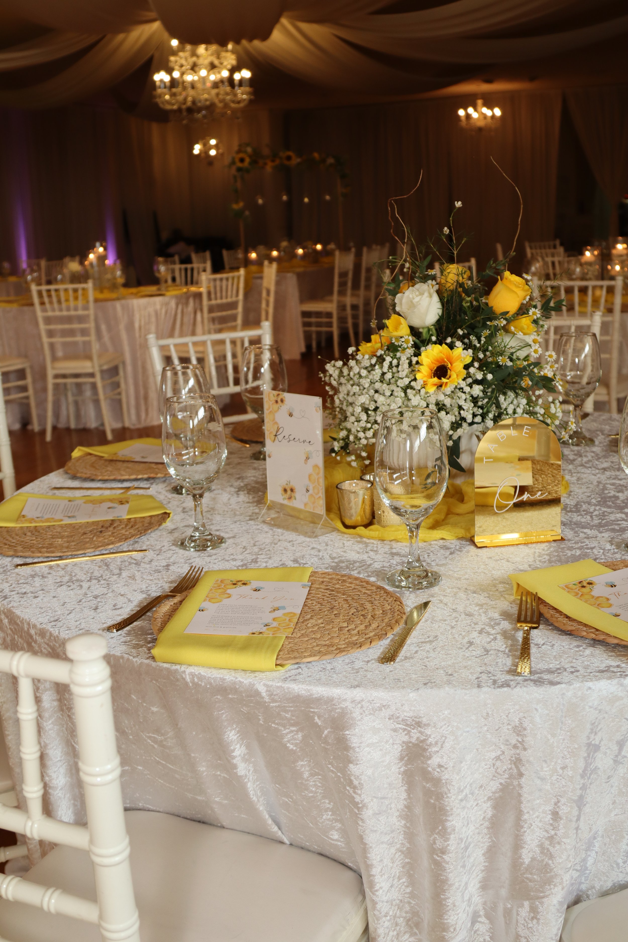A banquet table decorated with a floral centerpiece of yellow and white flowers, glassware, cutlery, yellow napkins, and a reservation card, set in an elegant event space with chandeliers and draped ceiling.