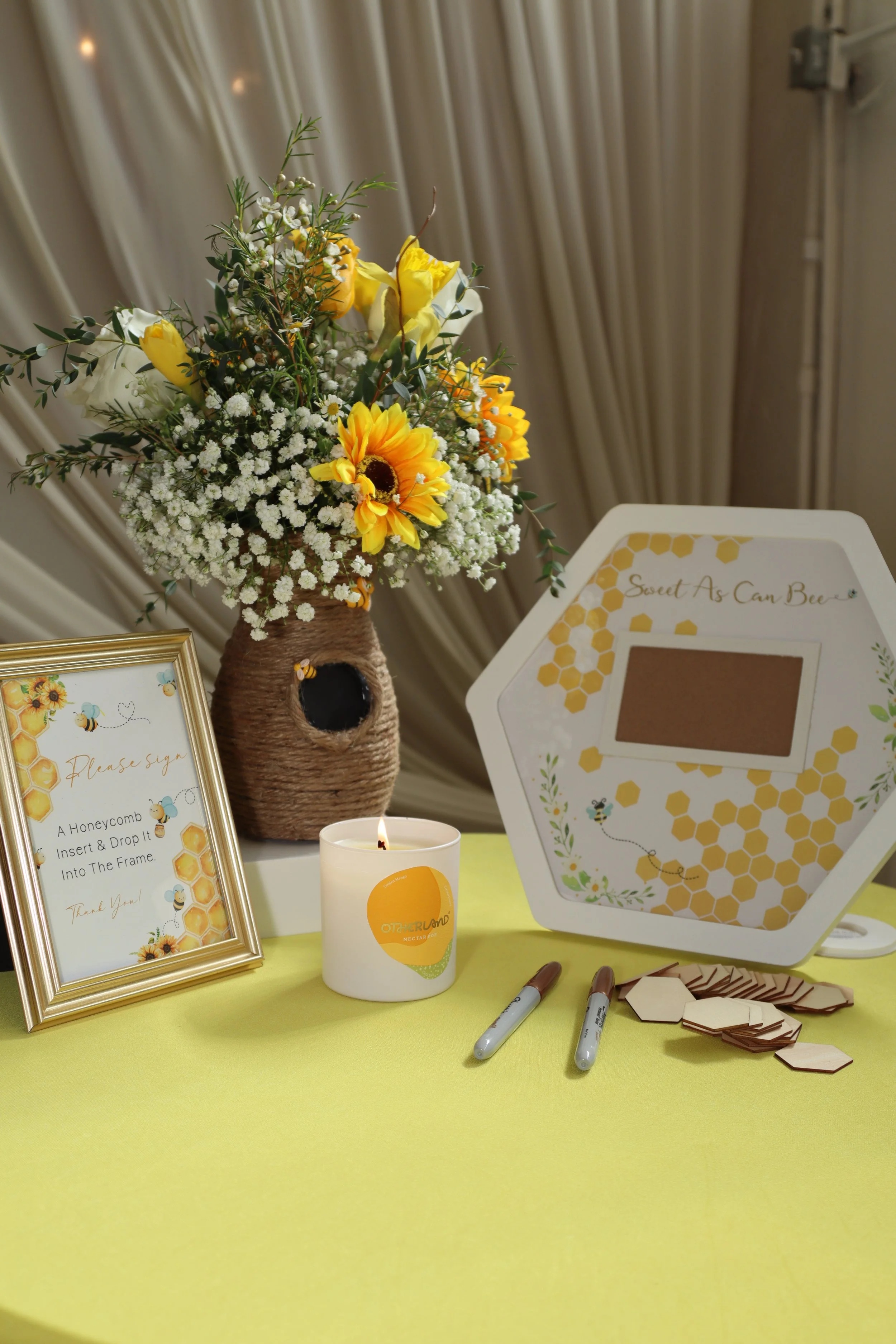A table with a large bouquet of yellow and white flowers in a woven vase, a framed sign with a honeycomb pattern and bee illustration, a white candle with an orange label, a honeycomb guestbook with a slot for inserting notes, and small hexagonal woo