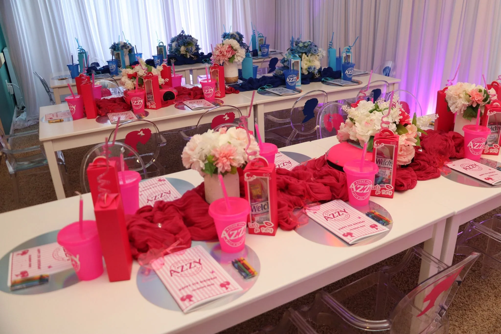 Decorated party tables with pink and blue themed balloons, flowers, drink cups, candies, and party favors in a room with white curtain backdrop.