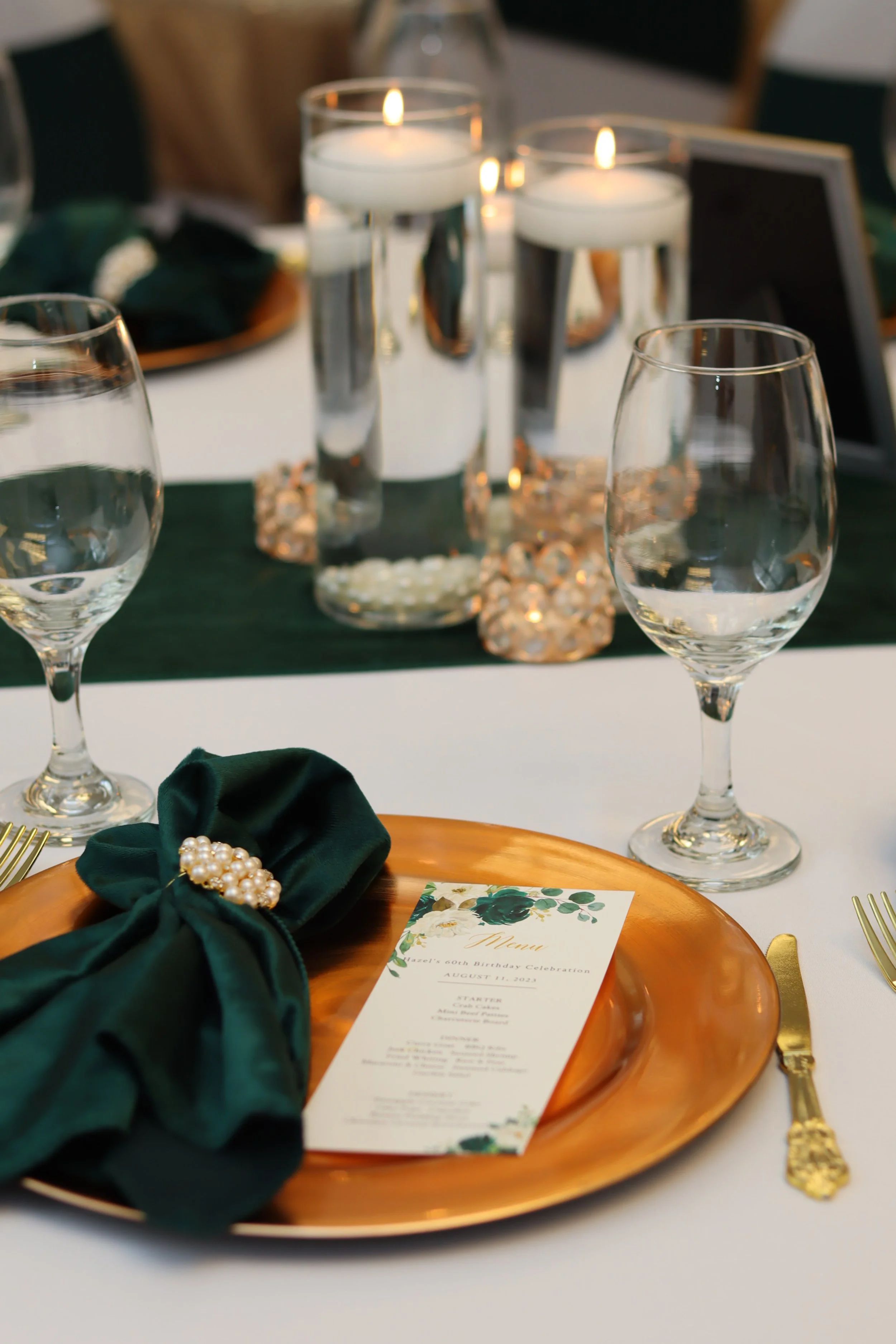 Table set for a celebration with a menu, gold flatware, green napkin with pearl brooch, water glasses, and candles