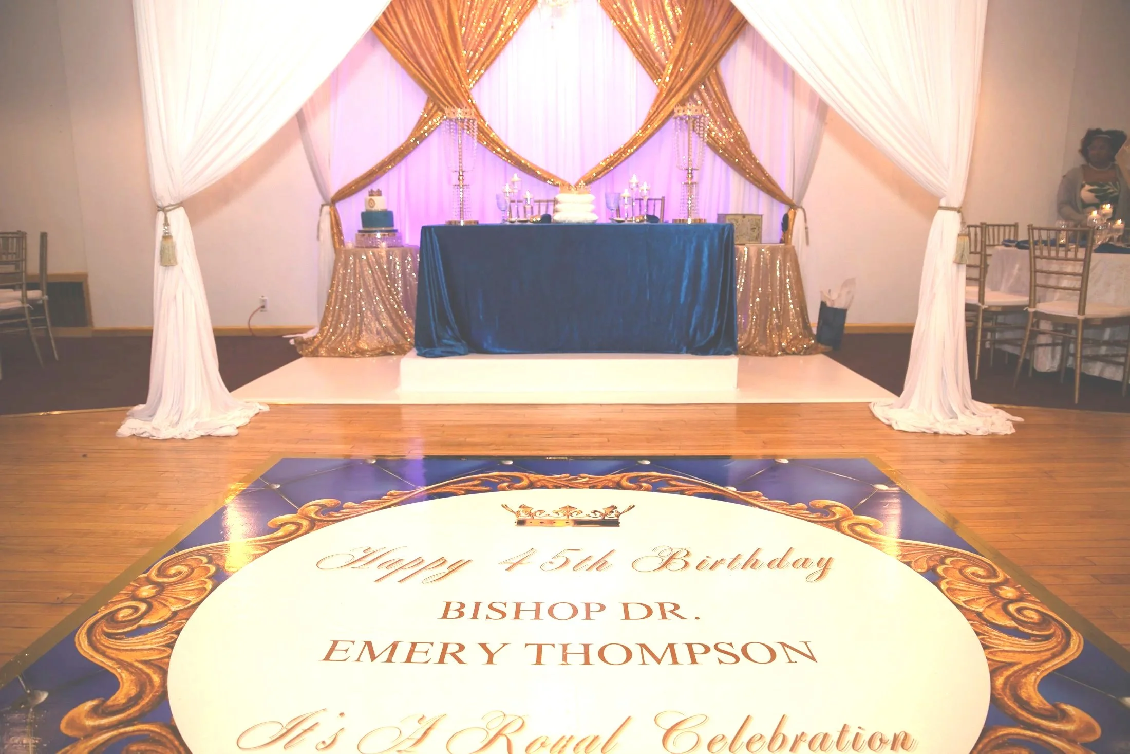 Decorative birthday celebration setup with a stage, gold and white draped curtains, a blue tablecloth-covered table with a cake, and a floor sign that reads 'Happy 45th Birthday Bishop Dr. Emery Thompson, His Royal Celebration'.