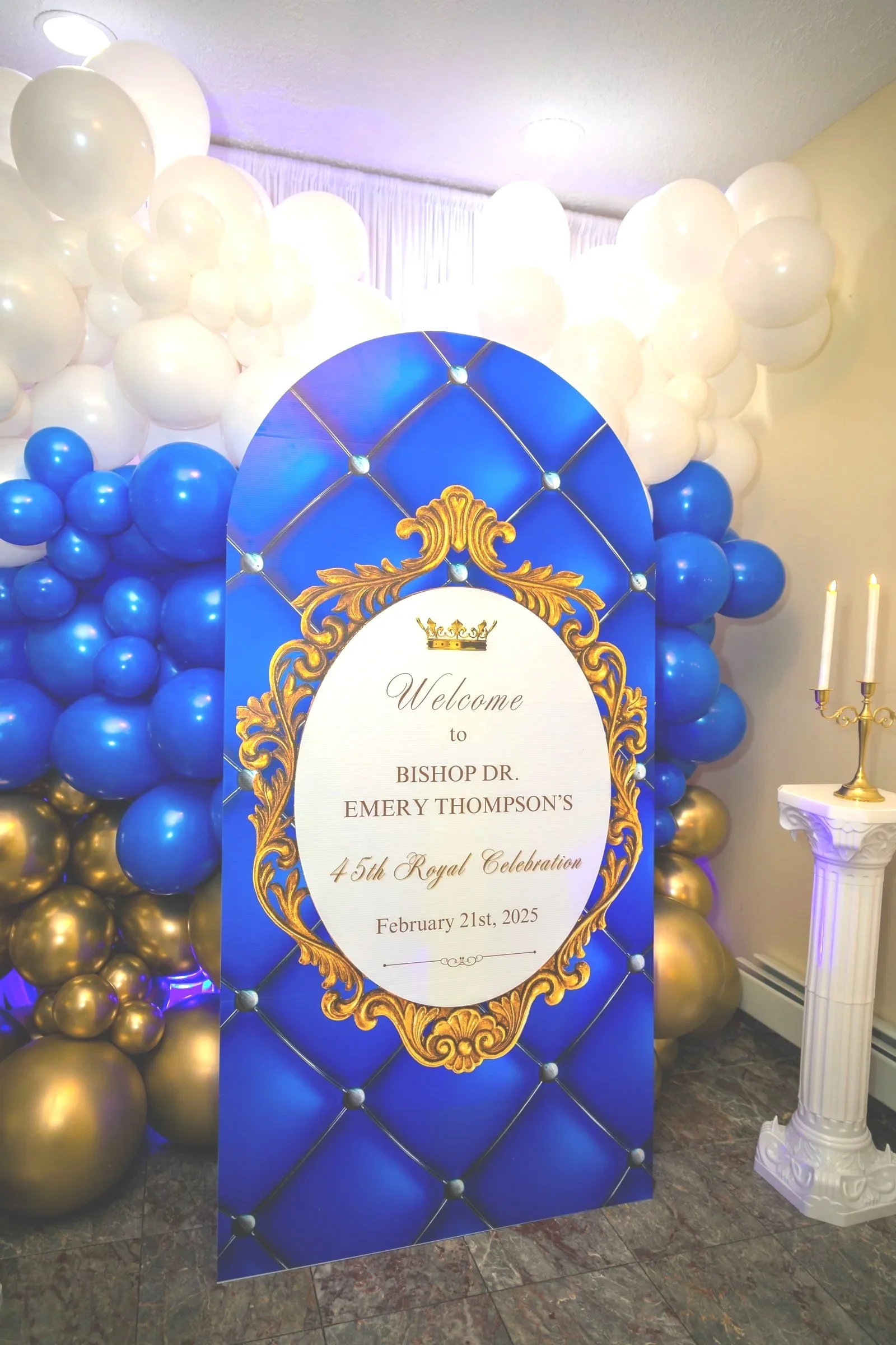 Large decorative welcome sign for Bishop Dr. Emery Thompson's 45th Royal Celebration on February 21st, 2025, with a backdrop of white, blue, and gold balloons and a white pedestal with a candelabra.