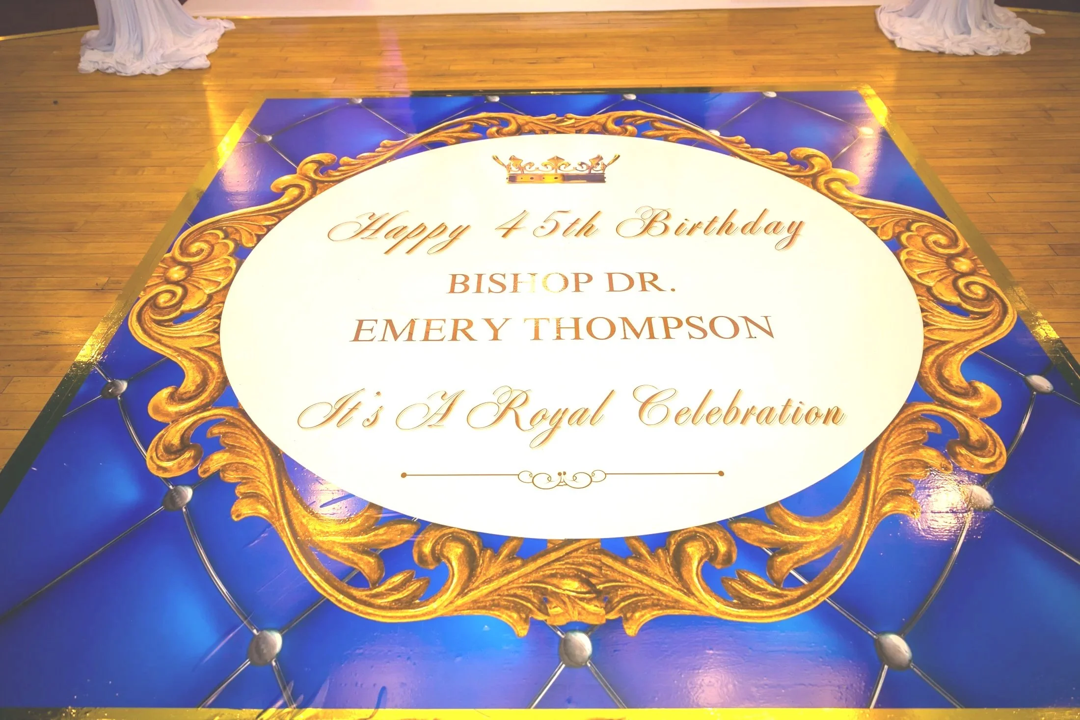 A large decorative floor banner celebrating the 45th birthday of Bishop Dr. Emery Thompson with gold ornate borders and text, on a hardwood floor.