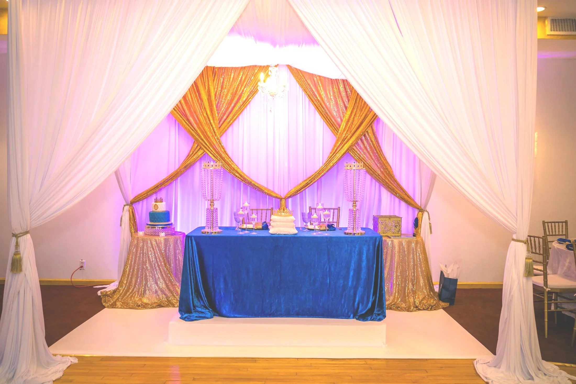 Decorated event table with blue tablecloth, gold drapes backdrop, pink lighting, cake, and candle holders, inside a white tent with draped curtains.