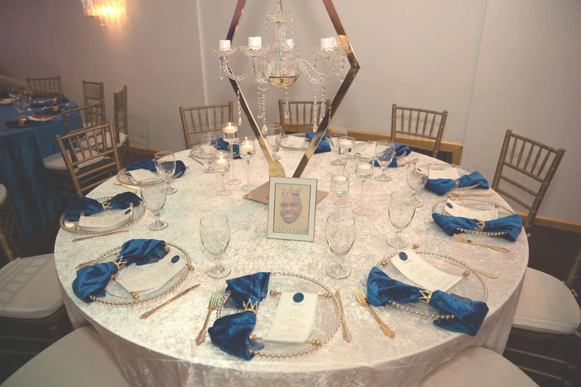 Round banquet table decorated with a white tablecloth and blue napkins, set with glassware, gold utensils, and a menu at each place setting, featuring a tall ornate crystal chandelier centerpiece and a reserved sign with a man's photo.