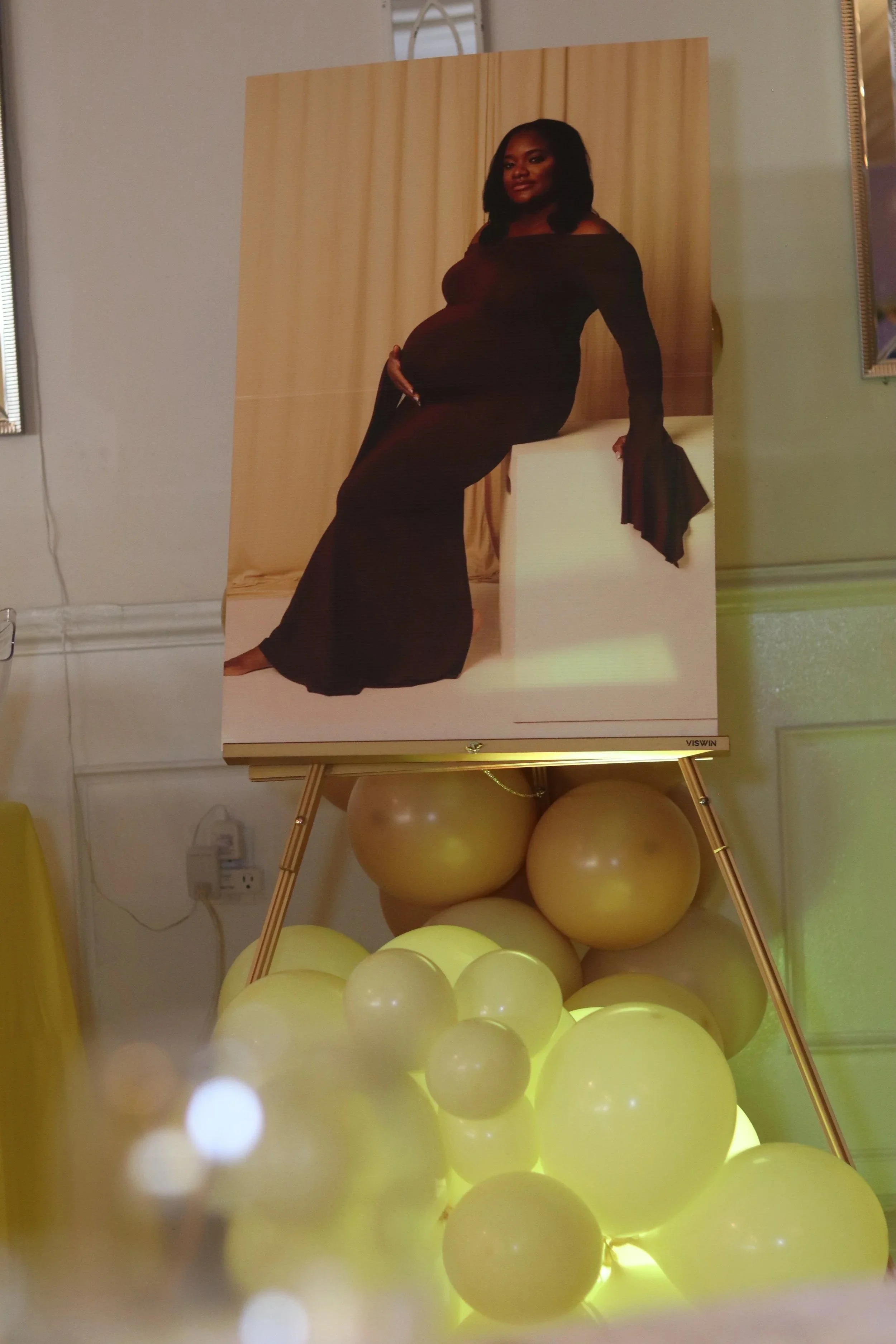 A portrait photograph of a pregnant woman wearing a long black dress, seated with her hand on her belly, displayed on a stand above a cluster of illuminated yellow balloons.