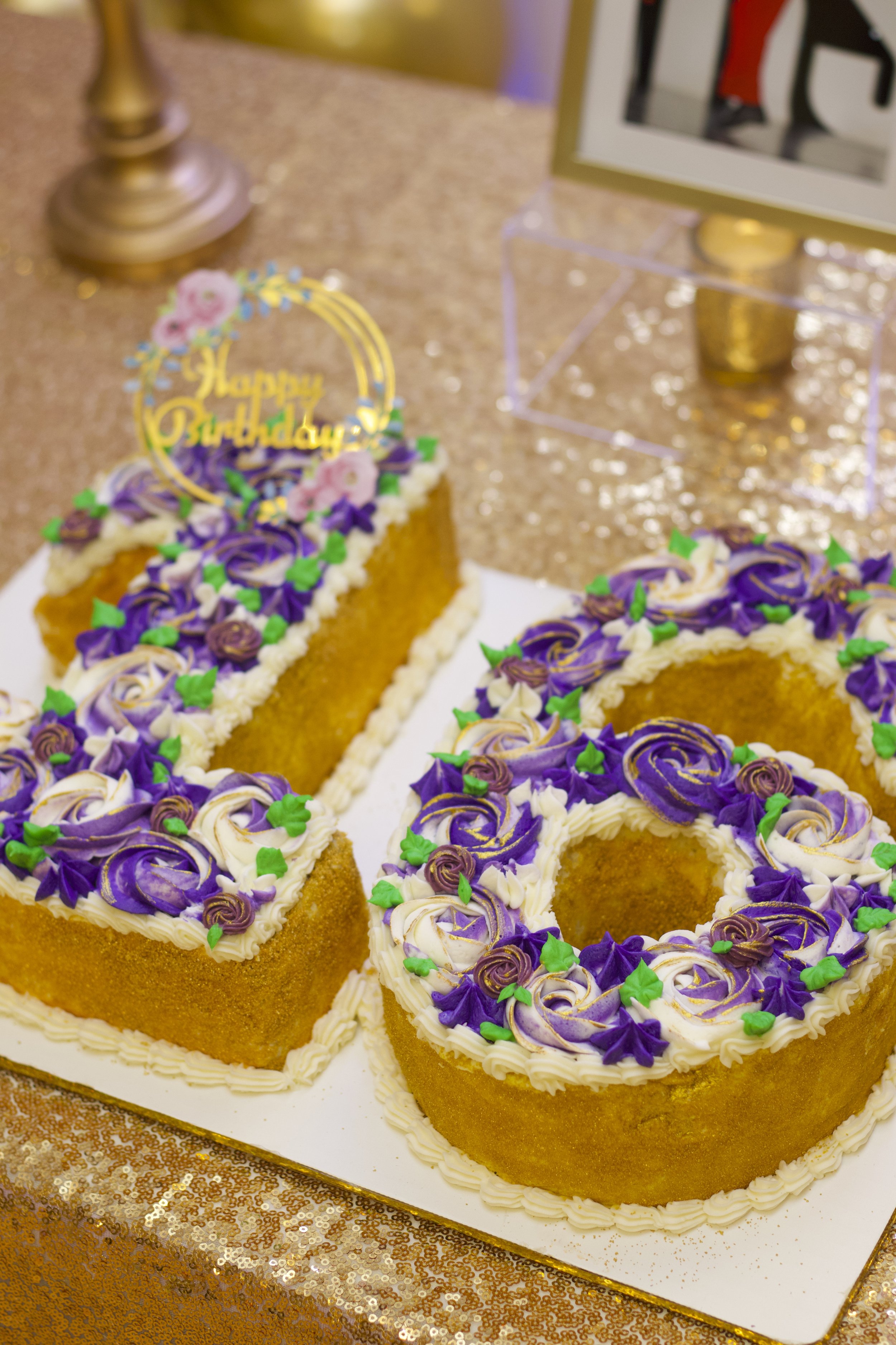 A birthday cake decorated with purple, white, and gold frosting roses, with a 'Happy Birthday' topper, on a decorated table.