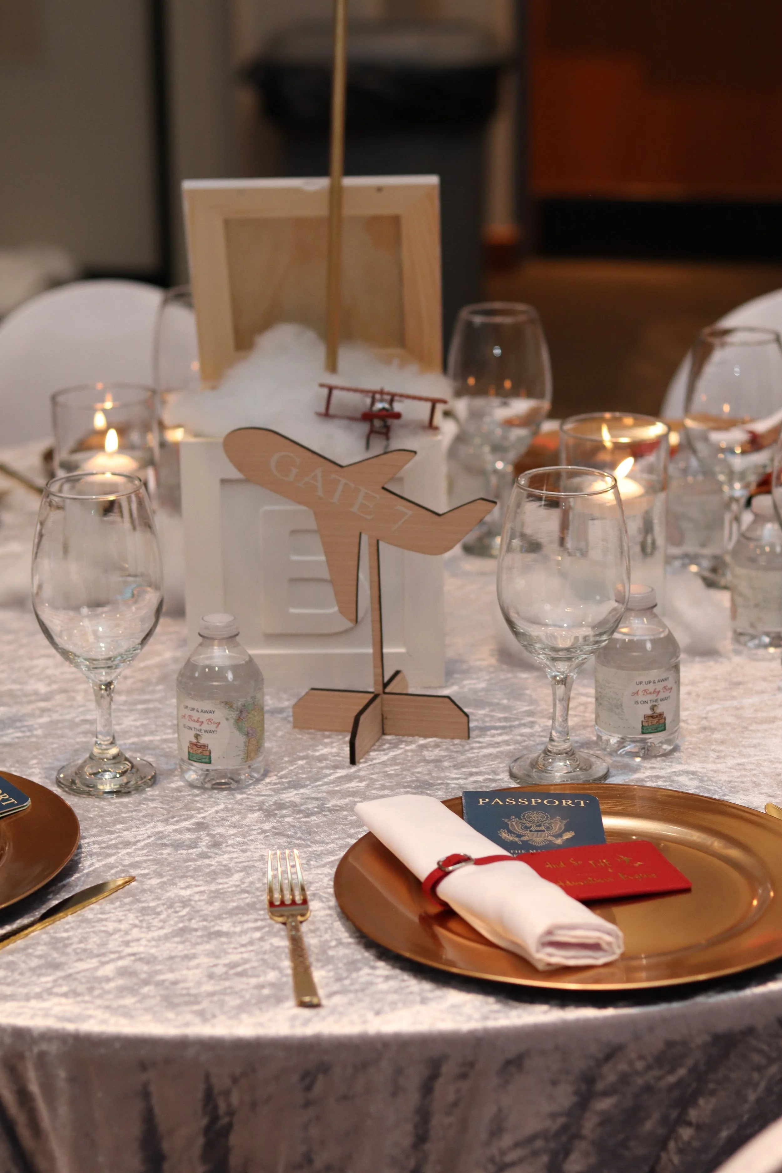 Table setting with gold charger plates, white napkins, gold utensils, glasses, water bottles, candles, and a wooden airplane and whale centerpieces, indicating a wedding or special event.