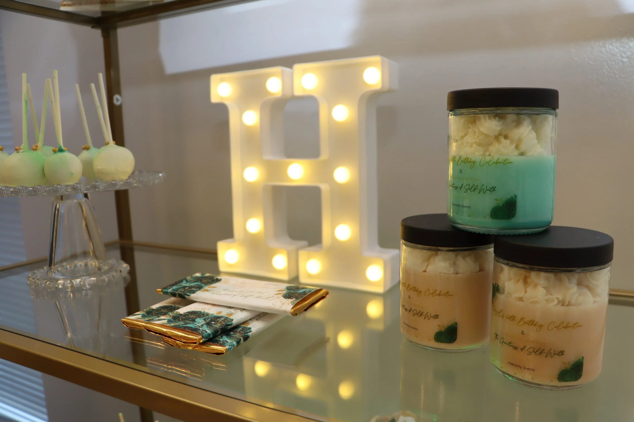 Decorative display featuring three jars of bath salts, a lit marquee letter "L", a tray of cake pops with green and white icing, and a set of wrapped gifts or party favors on a glass table.