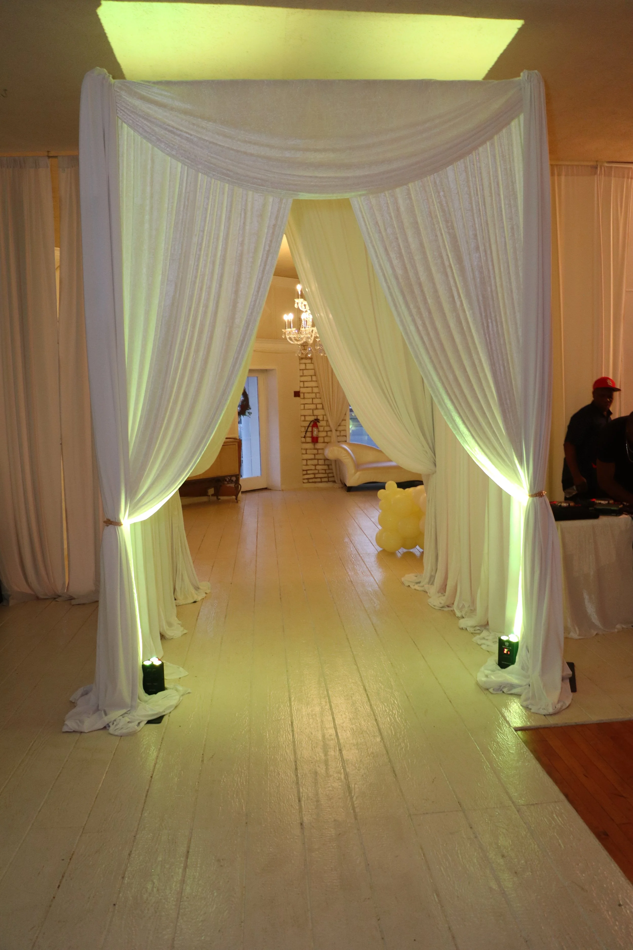 A decorated wedding or event entrance with white draped fabric and lighting, leading into a hall with a chandelier and seating area.