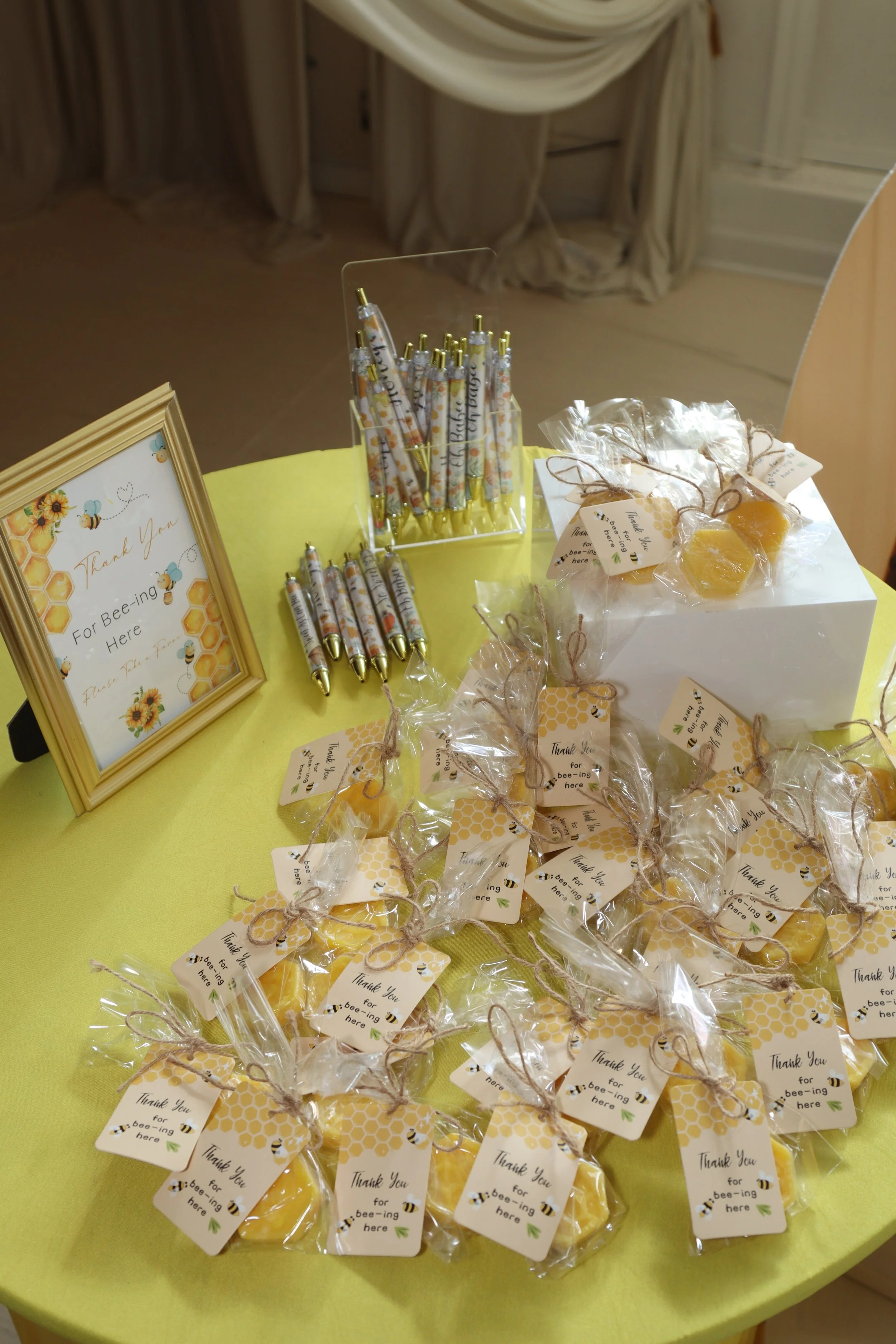 Table with thank you gifts for bee-ing here, including honey jars wrapped with thank you tags, bee-themed pens, and a framed sign with bee and honeycomb designs.