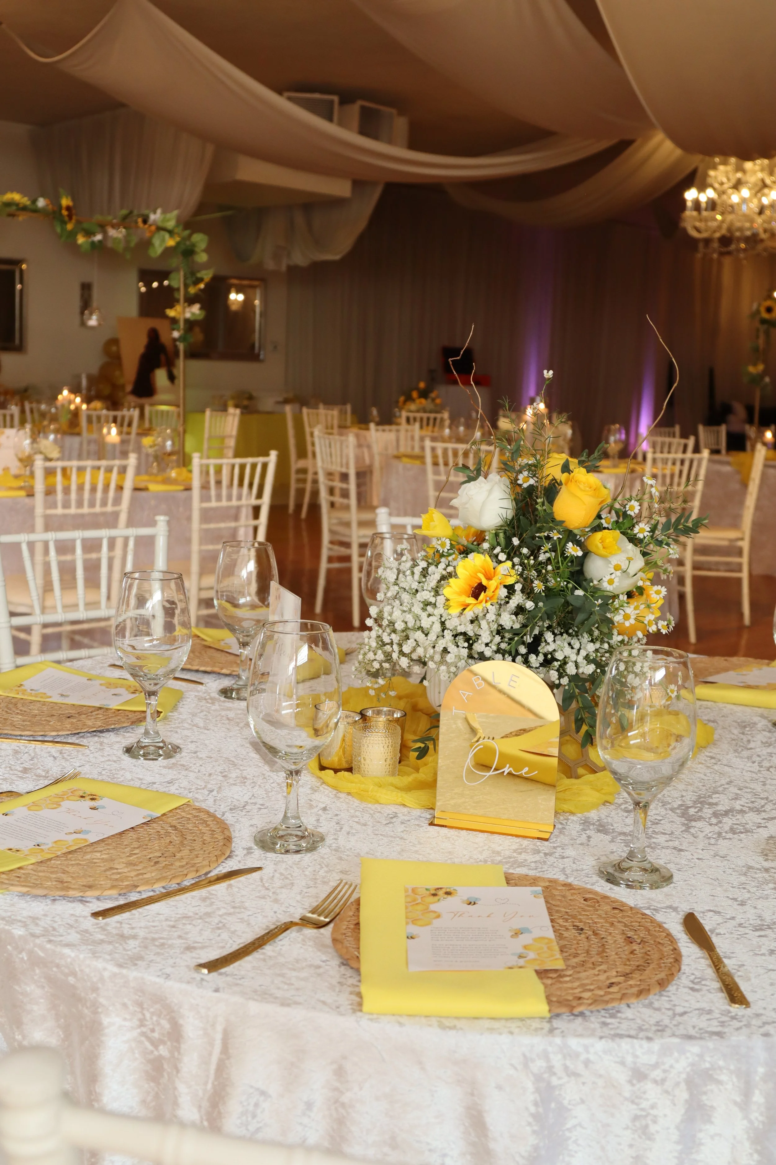 Wedding reception table decorated with yellow and white flowers, candles, glassware, gold cutlery, chairs, and a sign labeled 'Table One' with a menu.