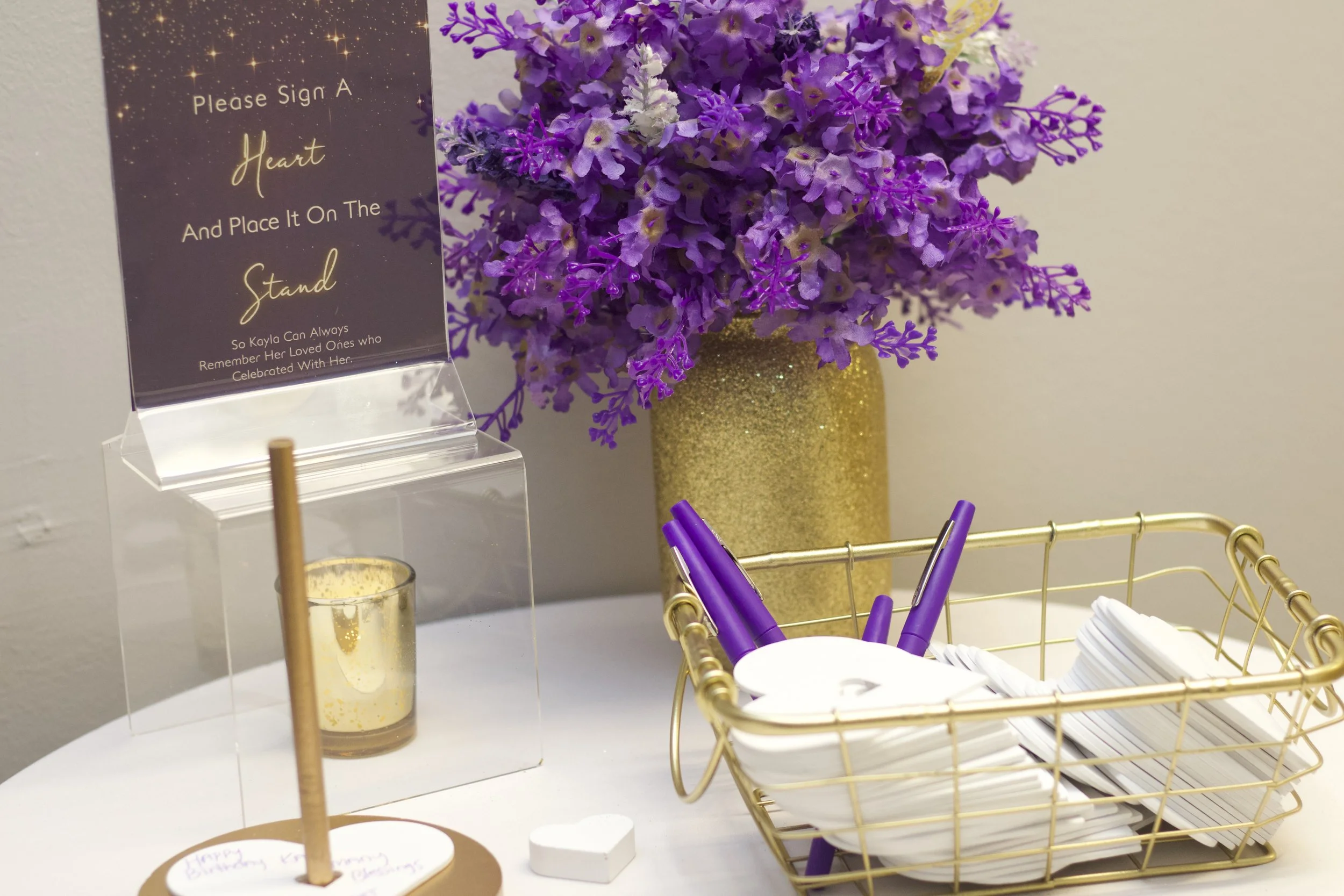 Table with purple and white artificial flowers in a gold vase, a clear acrylic stand with a sign that reads 'Please Sign A Heart and Place It on The Stand So Kayla Can Always Remember Her Loved Ones Who Celebrated With Her,' a small gold candle holde