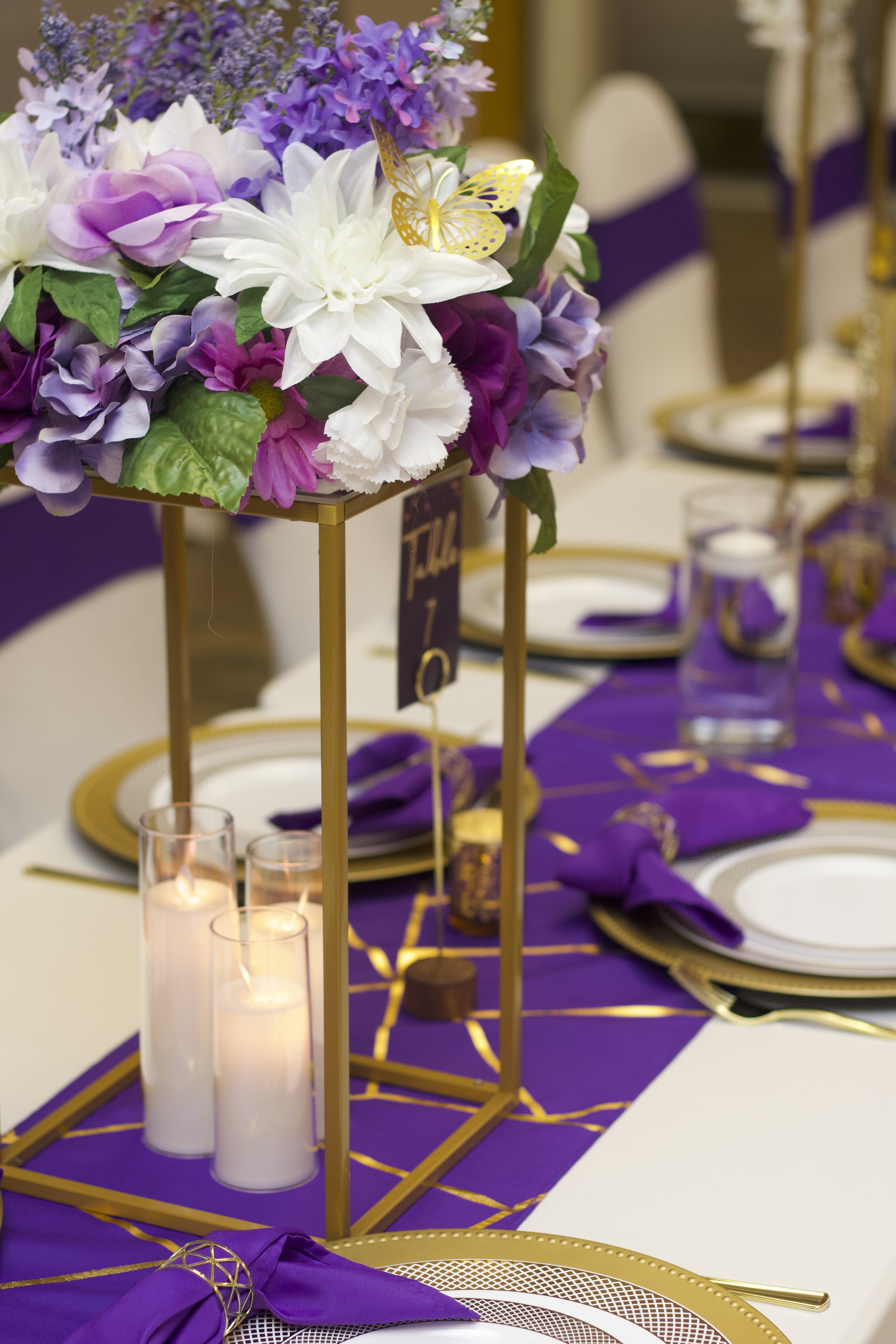 A table decorated for a formal event with a purple and gold color theme. It features a centerpiece with pink, white, purple flowers and yellow butterfly decorations, candles in glass holders, and tableware with purple napkins, gold-rimmed plates, and
