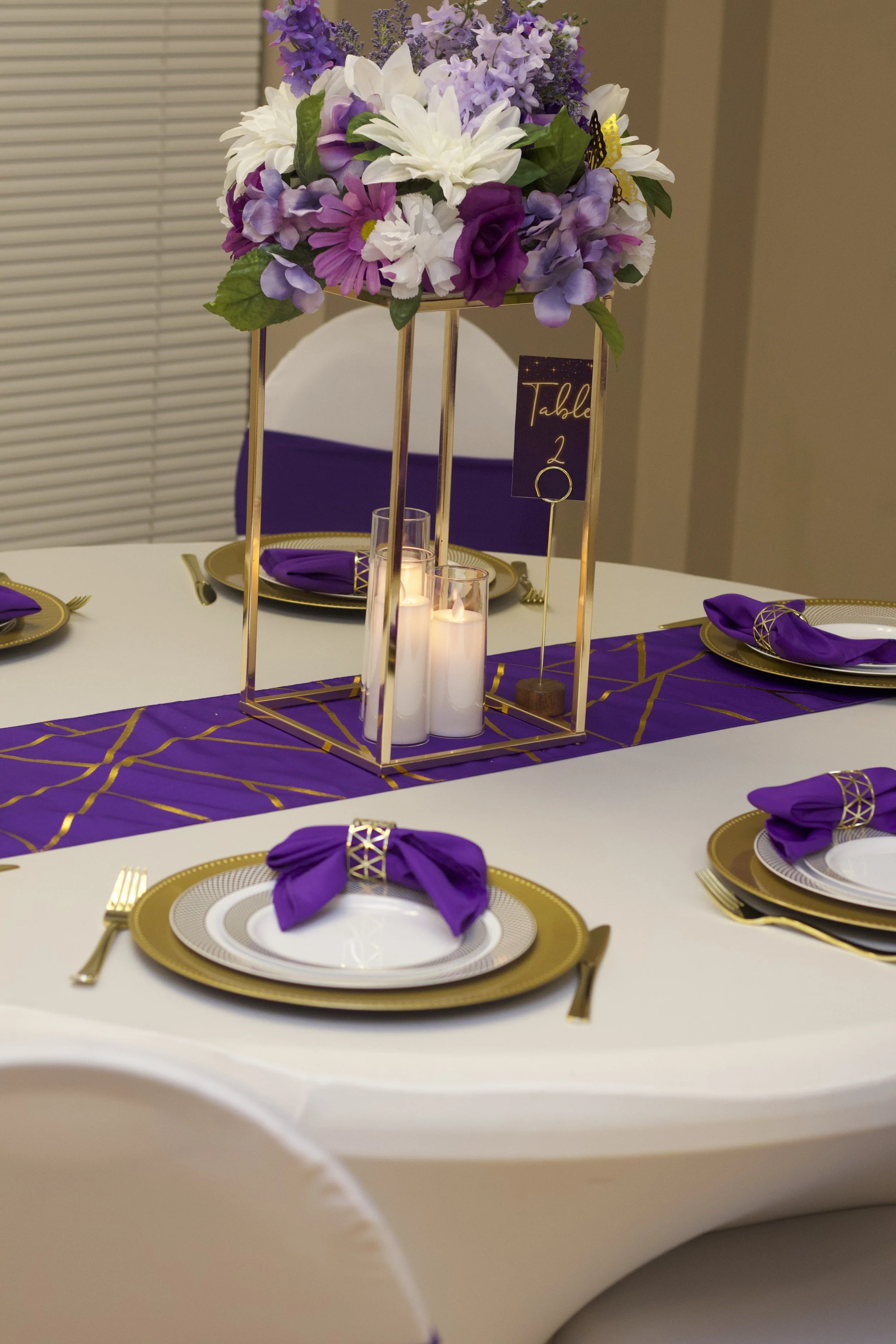 A table setting with purple and gold decorations, featuring white plates, purple napkins with gold rings, gold flatware, a purple table runner, a centerpiece with purple, white, and lavender flowers, candles, and a sign indicating "Table 2."