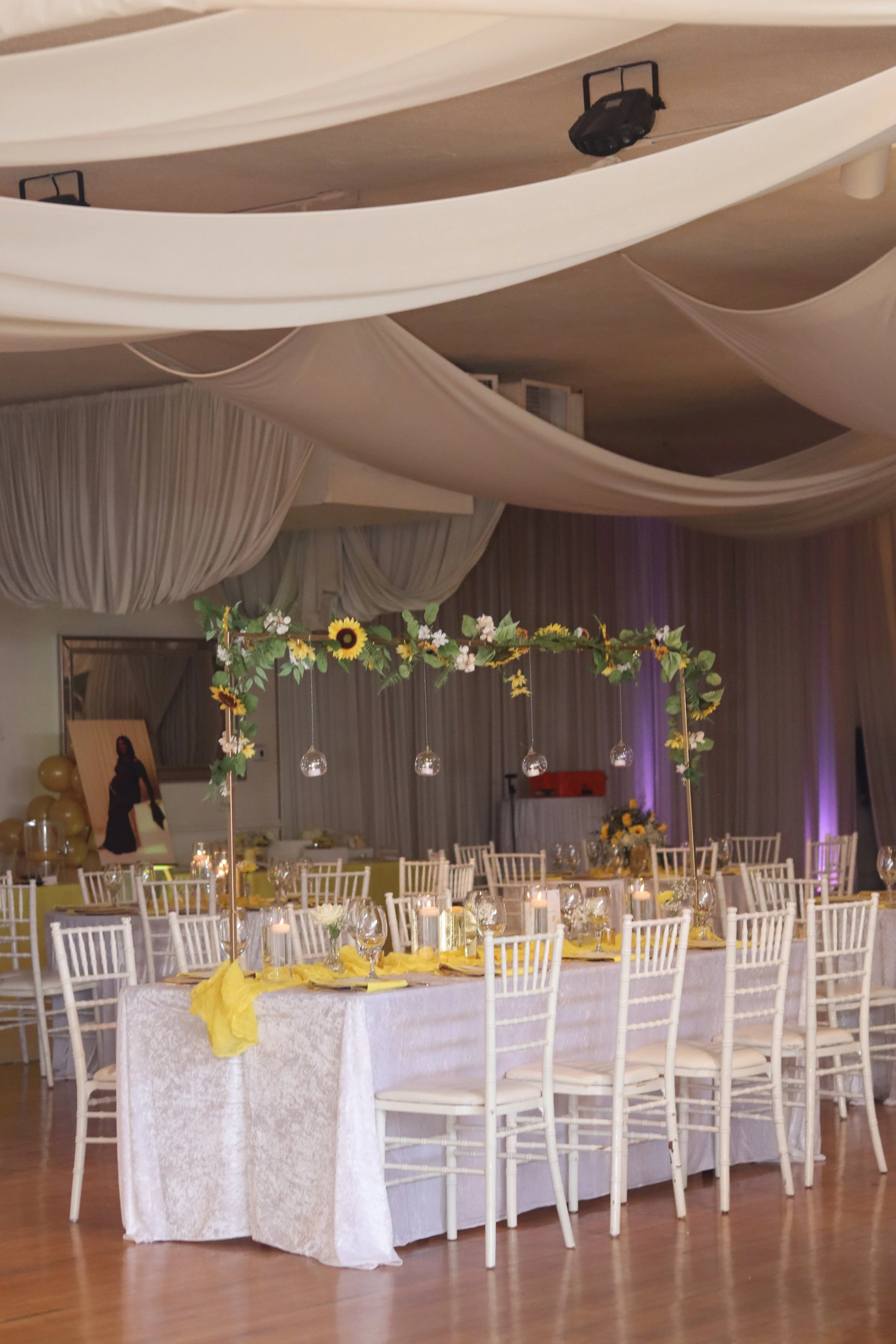 A decorated banquet table at a celebration with sunflowers and yellow accents, in a room with draped ceilings and purple uplighting.