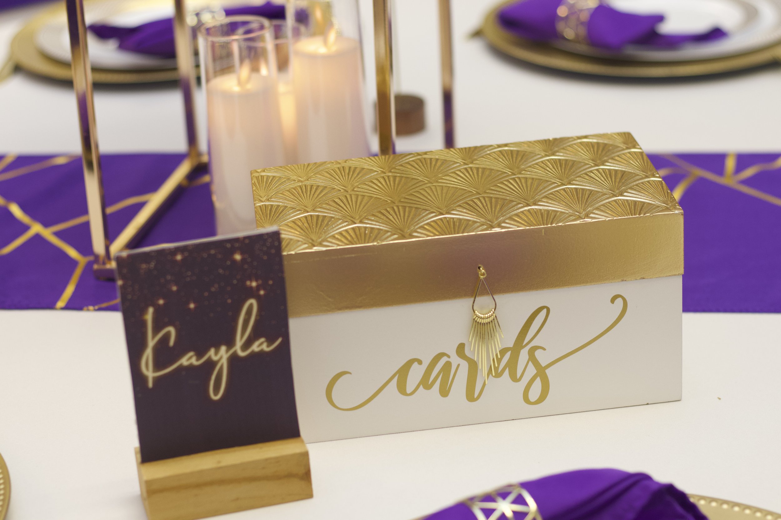 Table centerpiece with candles, a card box labeled 'cards' with a gold lid, and a place card with 'Kayla' written on it, decorated in gold, purple, and white.