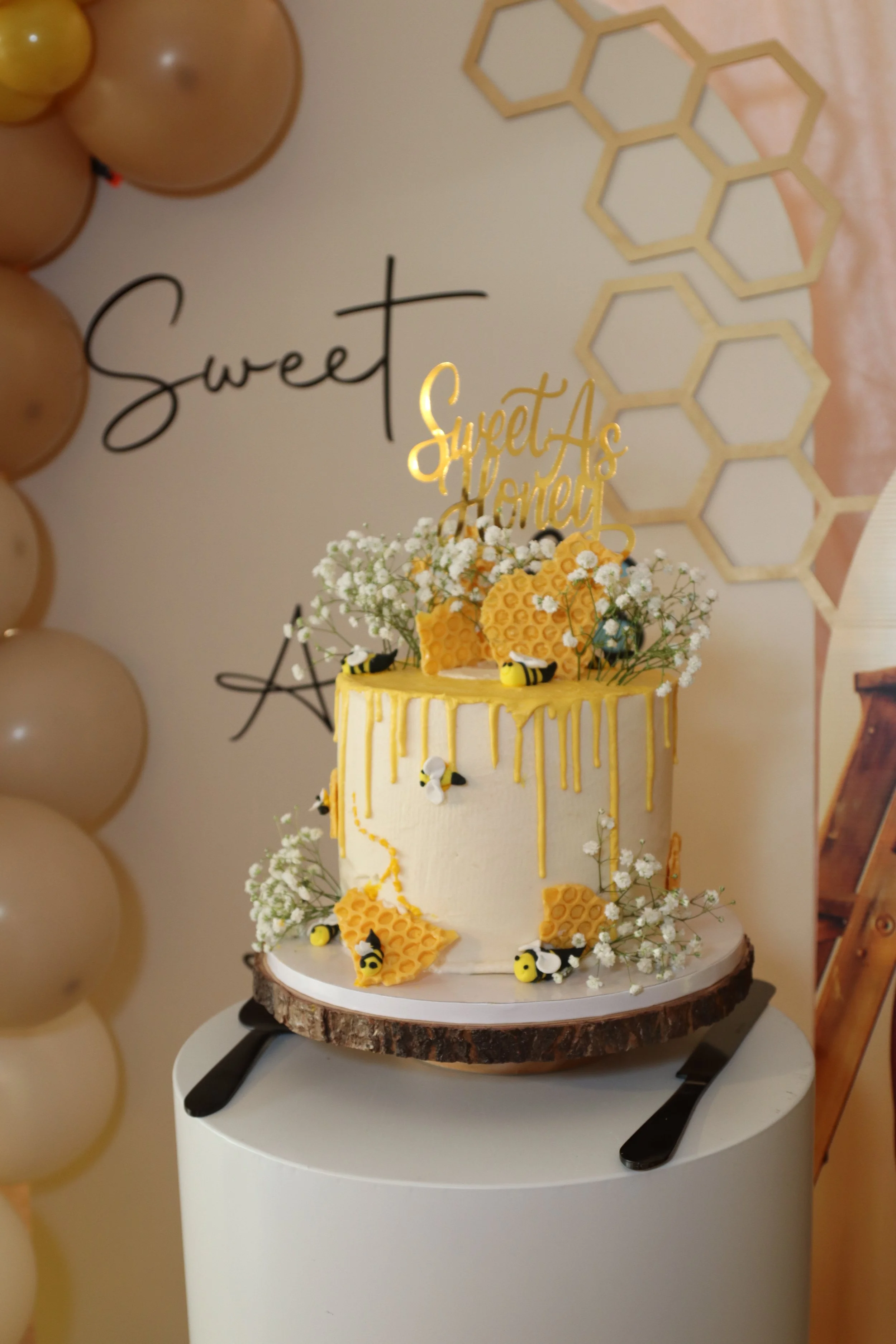 A honey-themed birthday cake decorated with honeycomb shapes, flowers, and small bees, placed on a tree slice cake stand, with a gold 'Sweet As Honey' cake topper, set against a Honeycomb and 'Sweet' sign background.