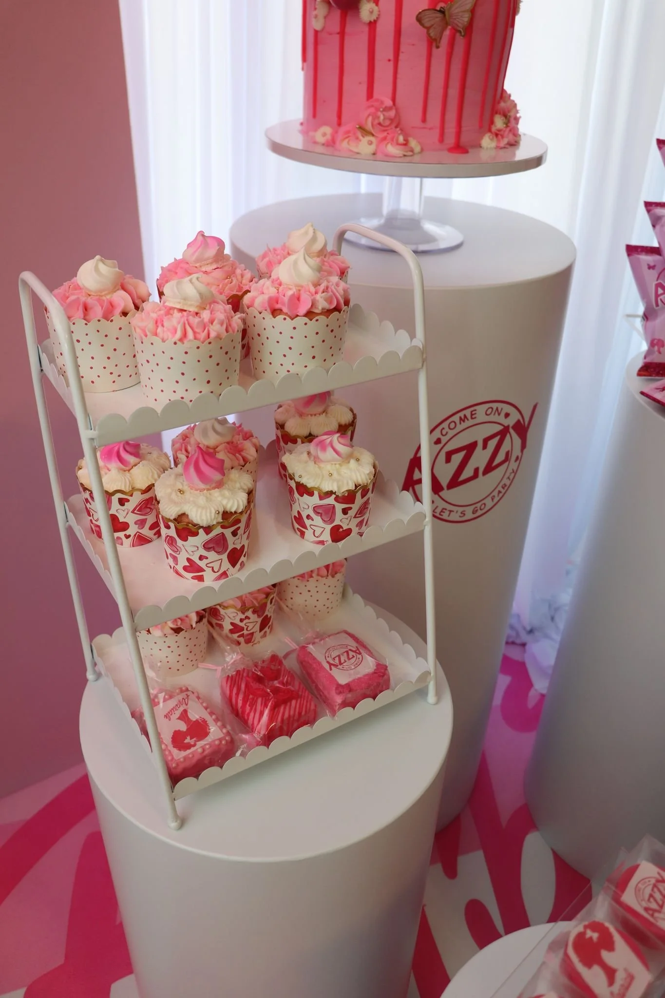 Display of Valentine-themed cupcakes and candies on a white stand with a pink cake on a taller white pedestal in the background, decorated with pink drips and flowers.