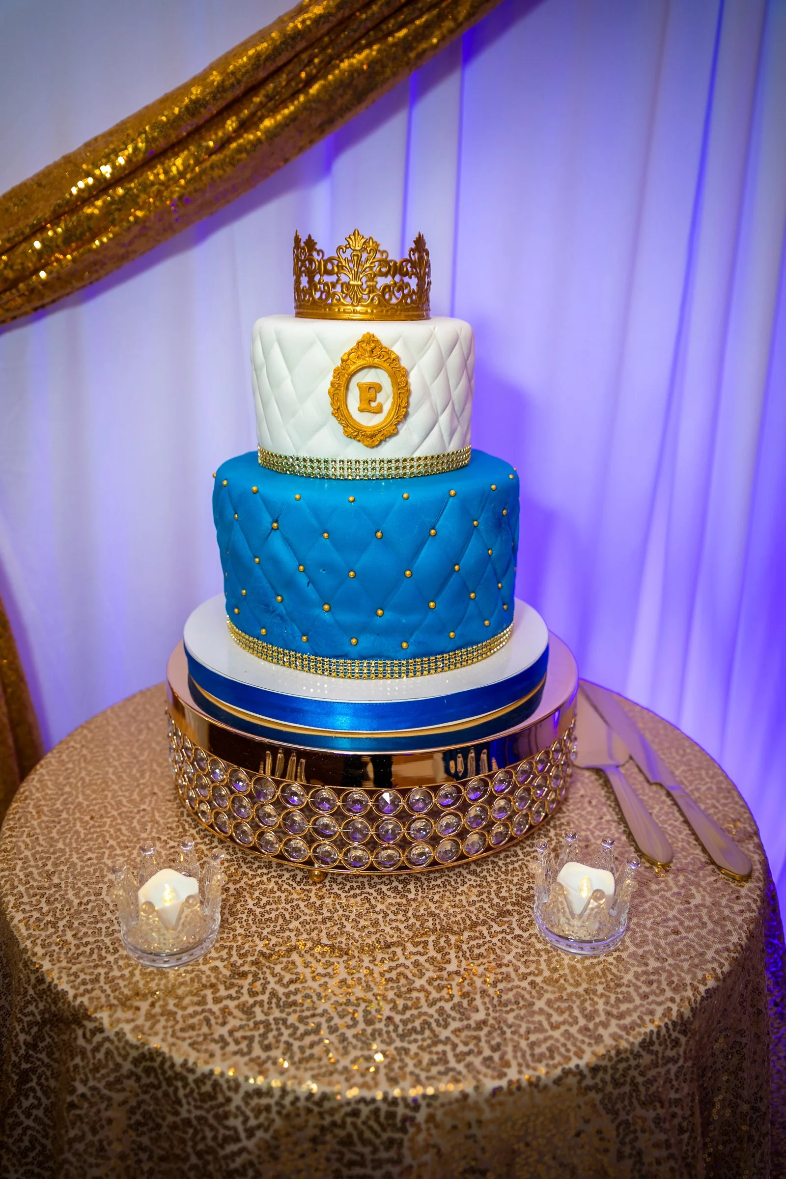 Three-tiered birthday cake with gold crown topper and monogram 'E' on white fondant, blue quilted fondant on the middle tier, decorated with gold beads; cake is on a gold sequined table with two candles and cake knives, and purple drapery in the back