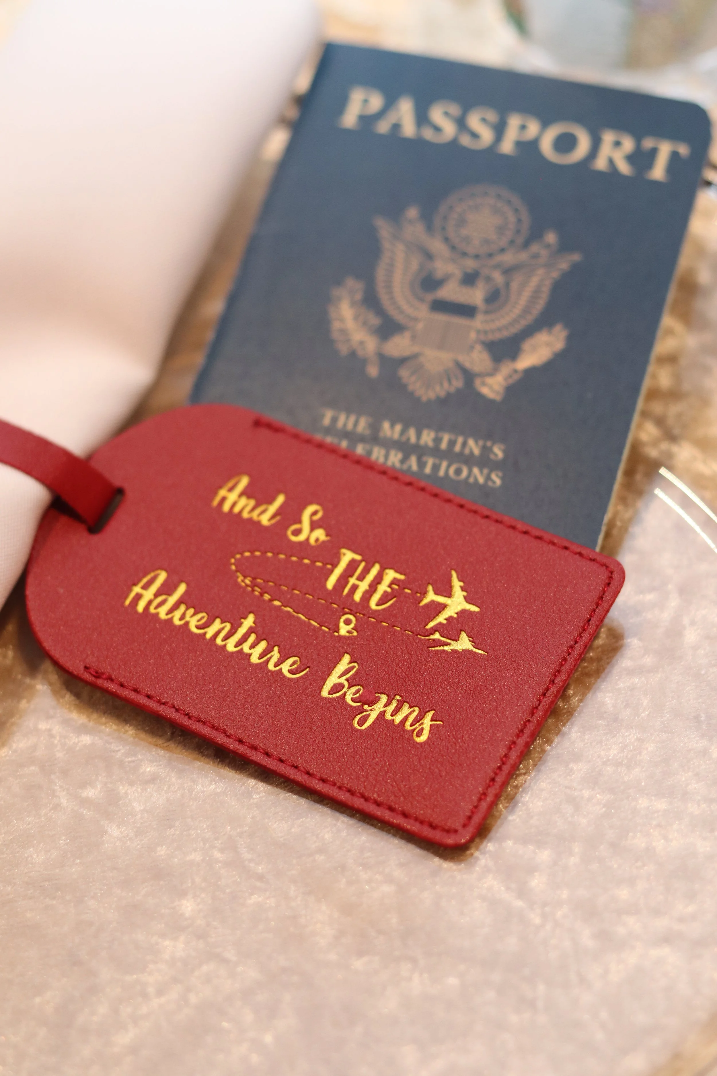 Close-up of a red travel tag with yellow text reading "And so the adventure begins" and a paper passport with a blue cover and gold emblem, placed on a shiny surface.