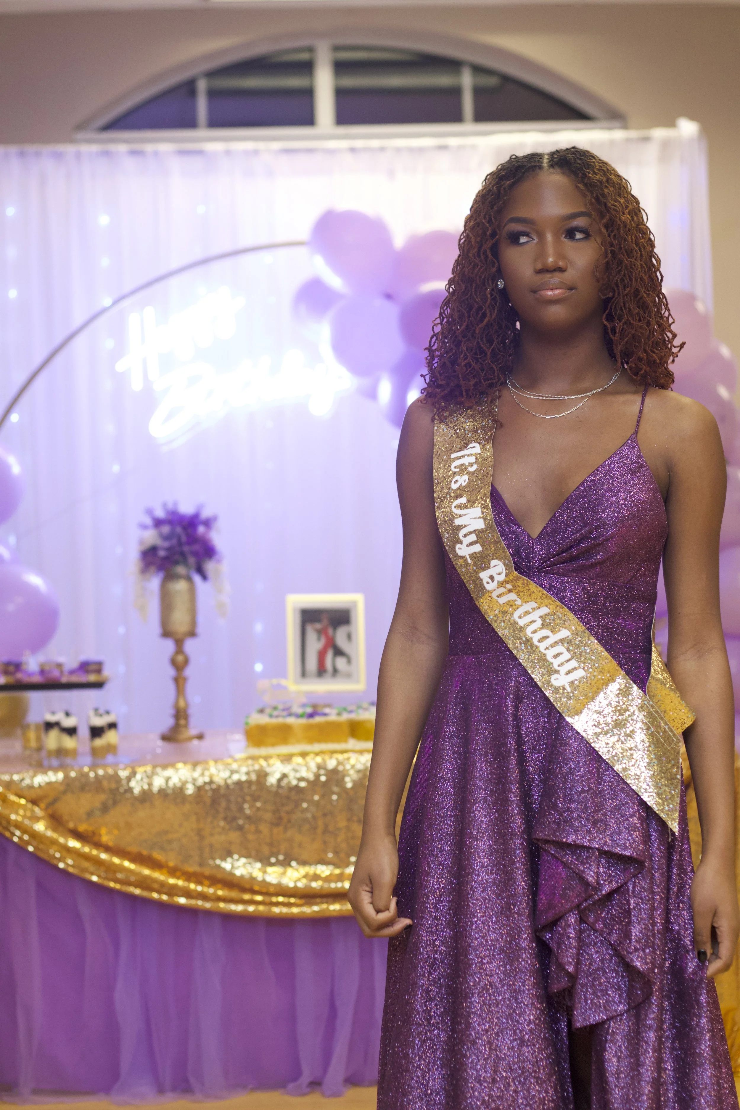 A young woman wearing a purple, glittery dress and a sash reading 'Tis My Birthday' at a birthday celebration with purple and gold decorations, balloons, and a cake in the background.