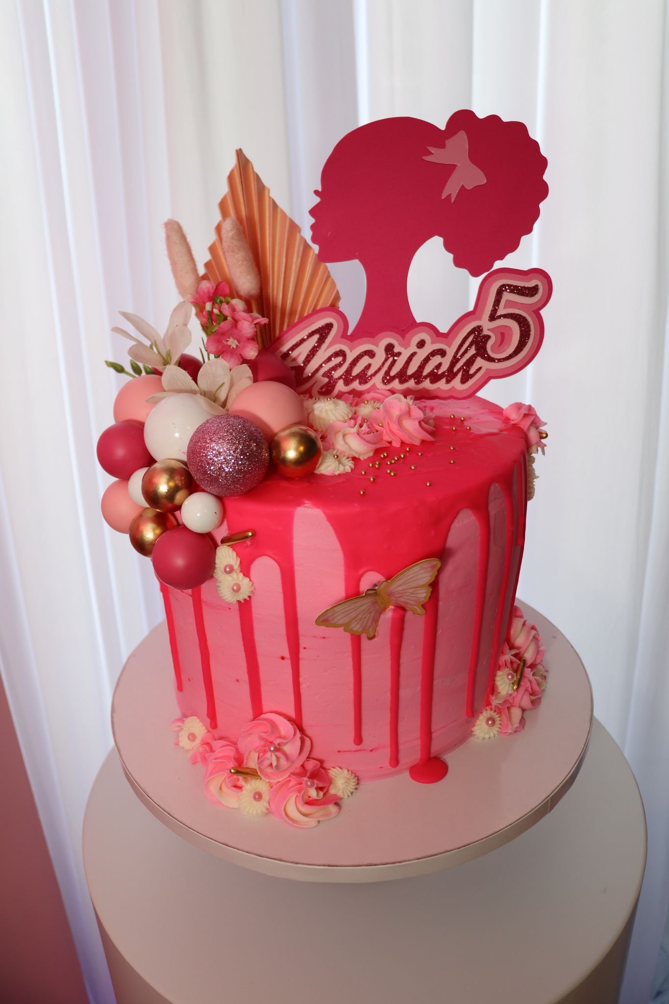 A pink birthday cake with a drip design, decorated with pink, white, and gold ornaments, flowers, and a silhouette topper of a girl's head with a bow and the name Azariah and the number 5.