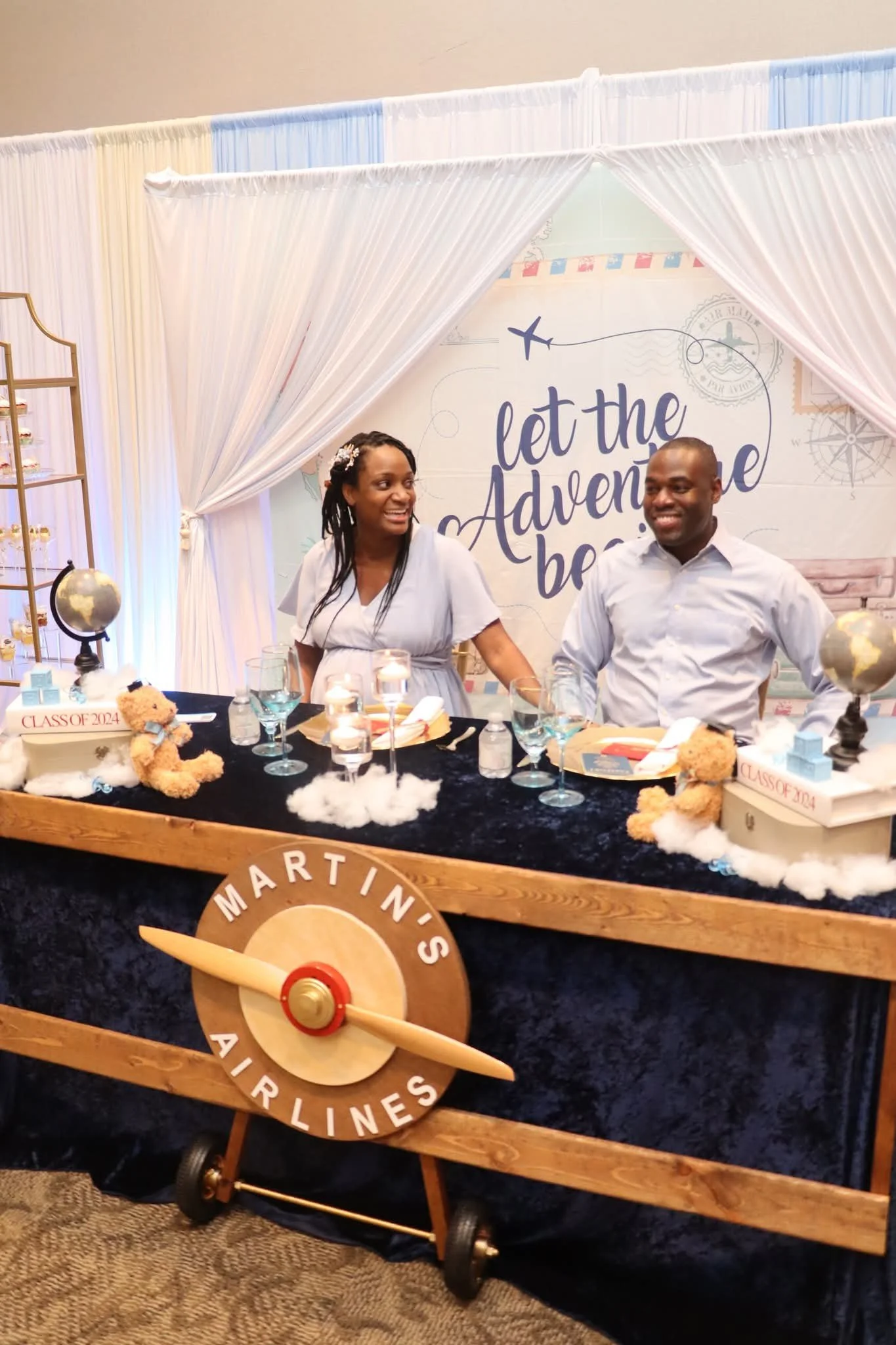 A couple sitting at a decorated table celebrating a graduation, with a backdrop that says 'let the adventure begin' and framed with white curtains, featuring travel-themed decor and a sign that says 'Martini's Airlines'.