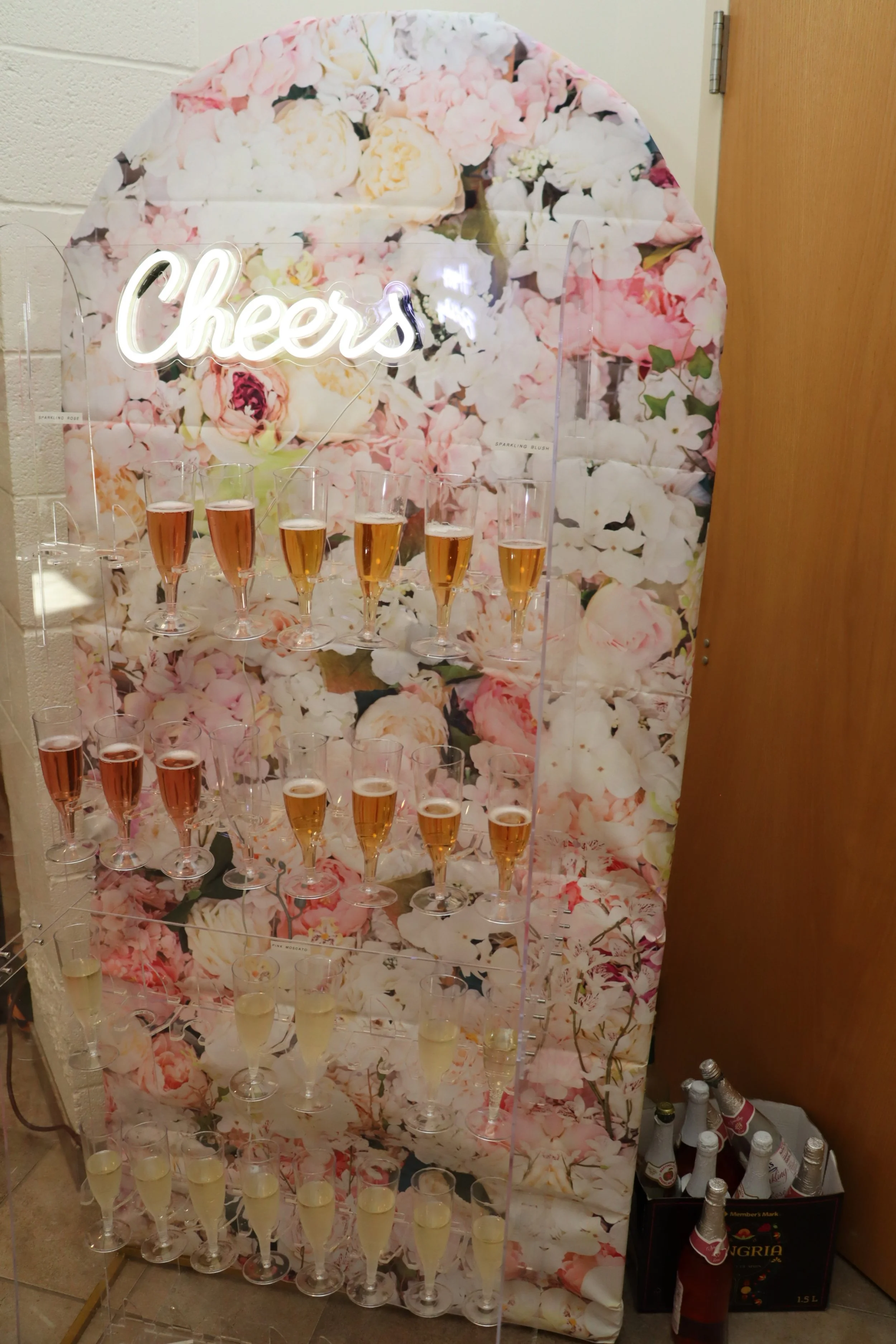 A champagne display with multiple tiers of champagne glasses filled with different colors of sparkling wine, set against a floral background with a neon sign that says 'Cheers'.