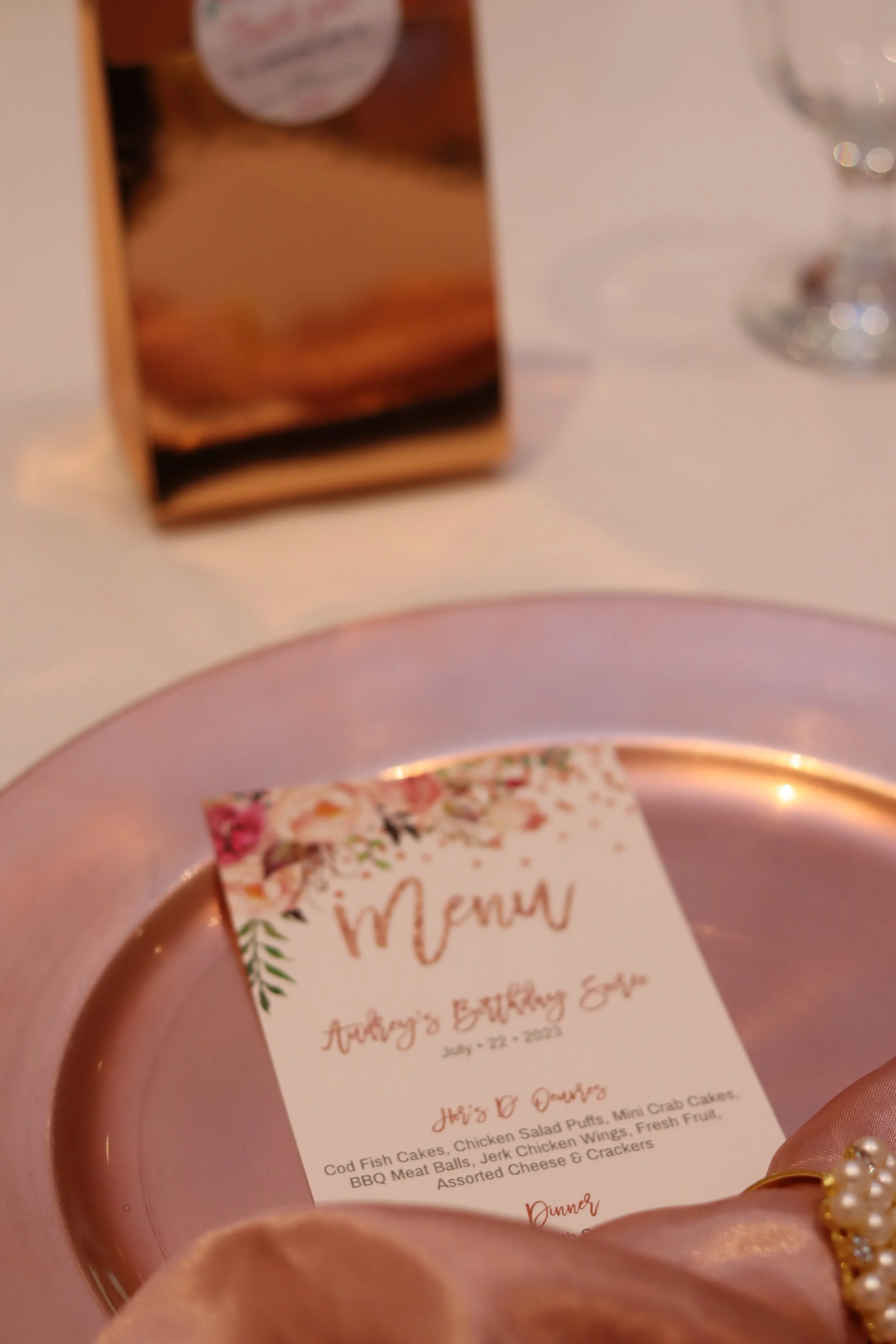 Table setting with a pink plate holding a floral-themed retirement party menu, a gold ring with pearls, and a brown gift box in the background.
