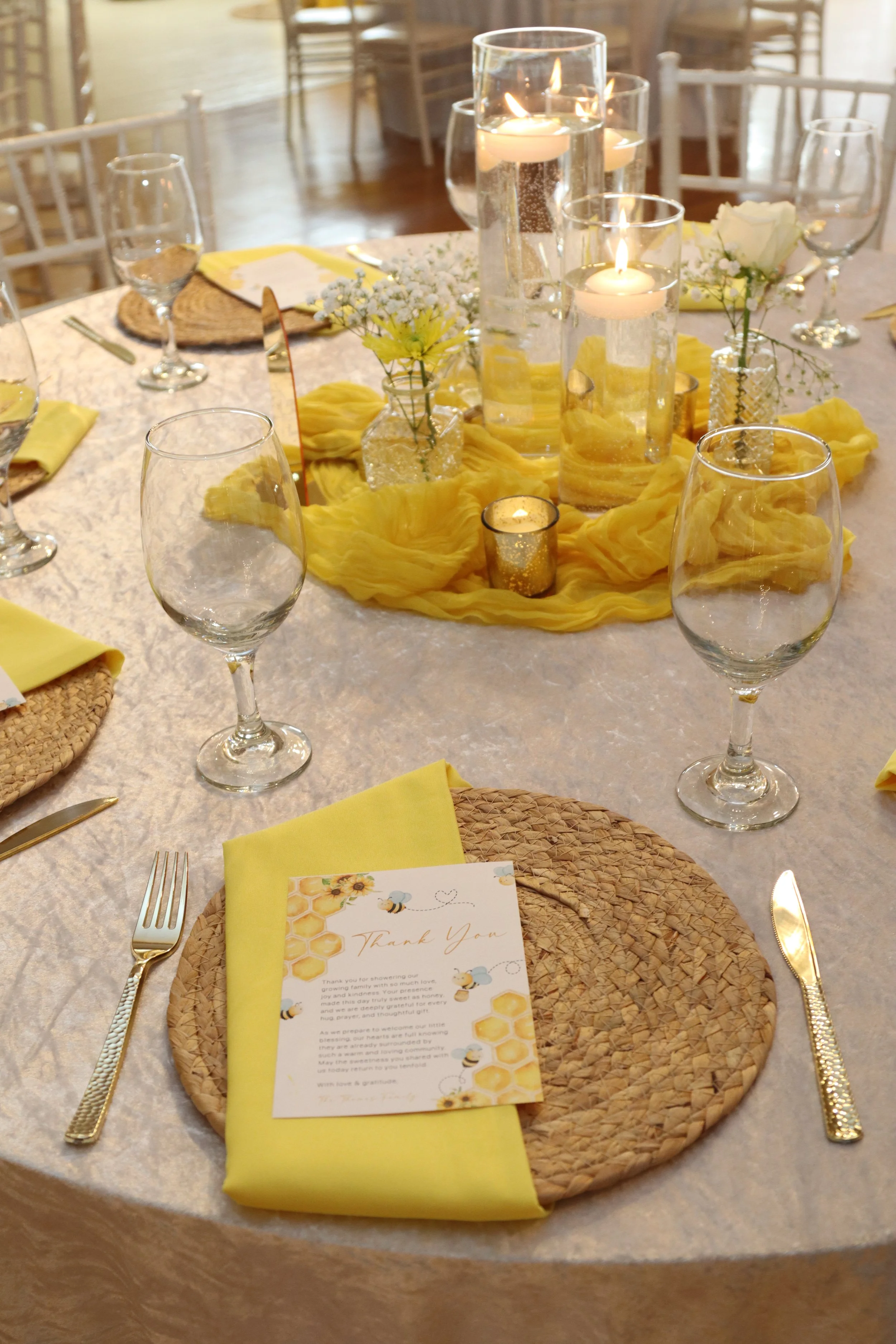A round dining table set for a celebration with a white tablecloth, yellow napkins, and woven placemats. The centerpiece features lit candles and small vases with white flowers and baby's breath, all draped with yellow fabric. There are wine glasses,