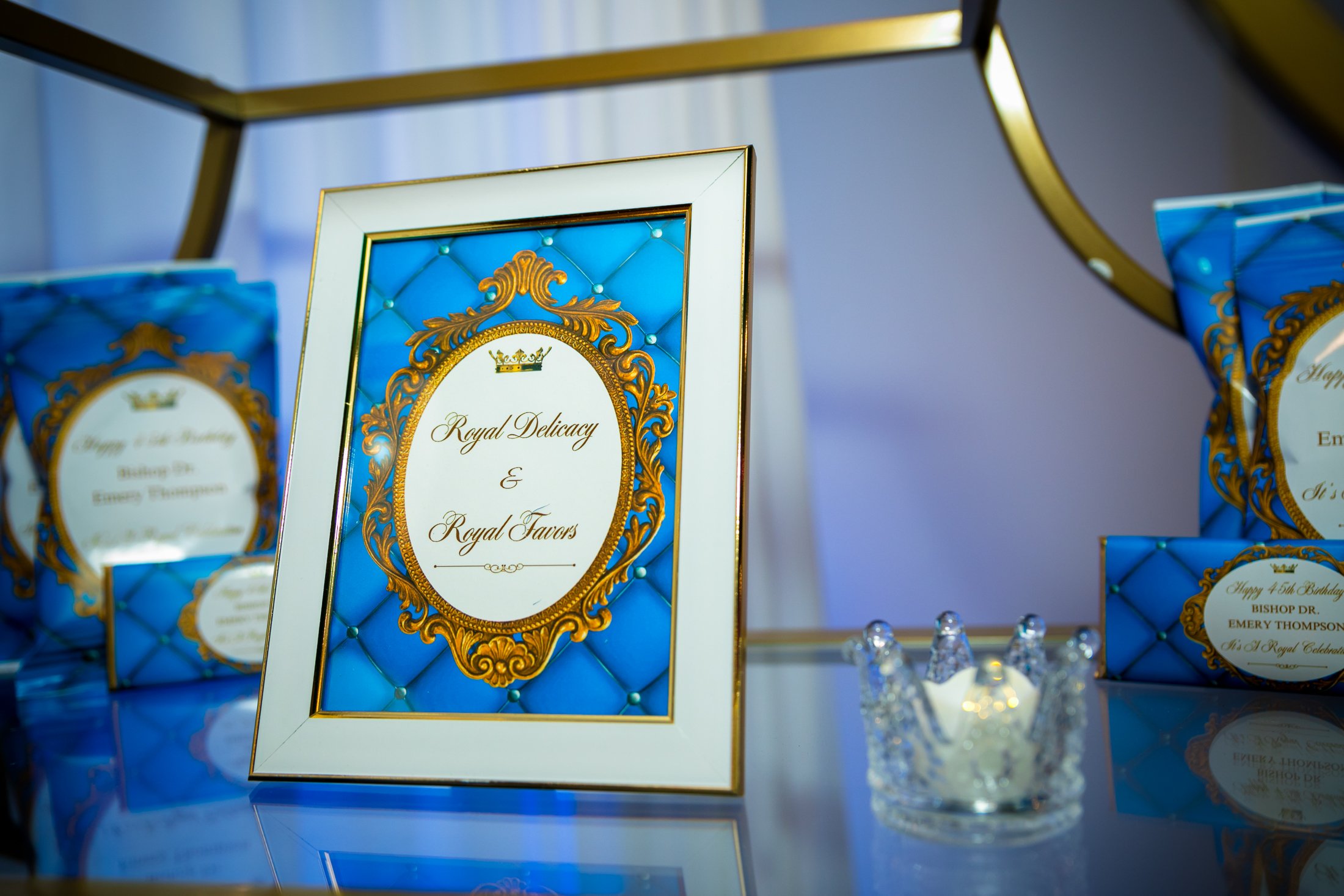 Decorative framed sign with gold and blue design, reading "Royal Delicacy & Royal Flavors," with smaller similar signs and a crystal crown-shaped candle holder on a reflective surface.