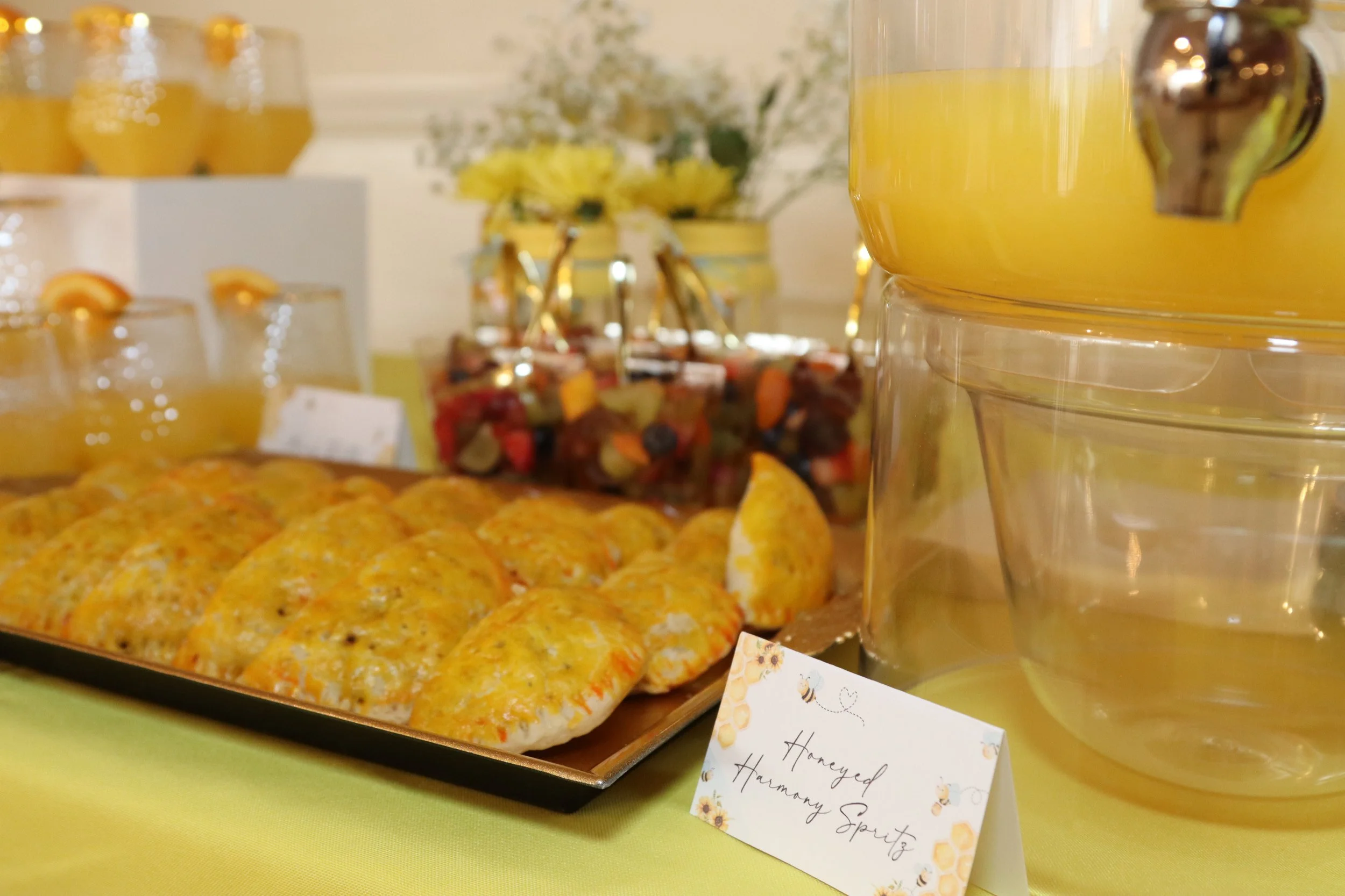 A tray of freshly baked honeyed cheese scones on a table with a decorative label, glass jars of honey and a honey fountain, a bowl of mixed fruit, and glasses of juice in the background.