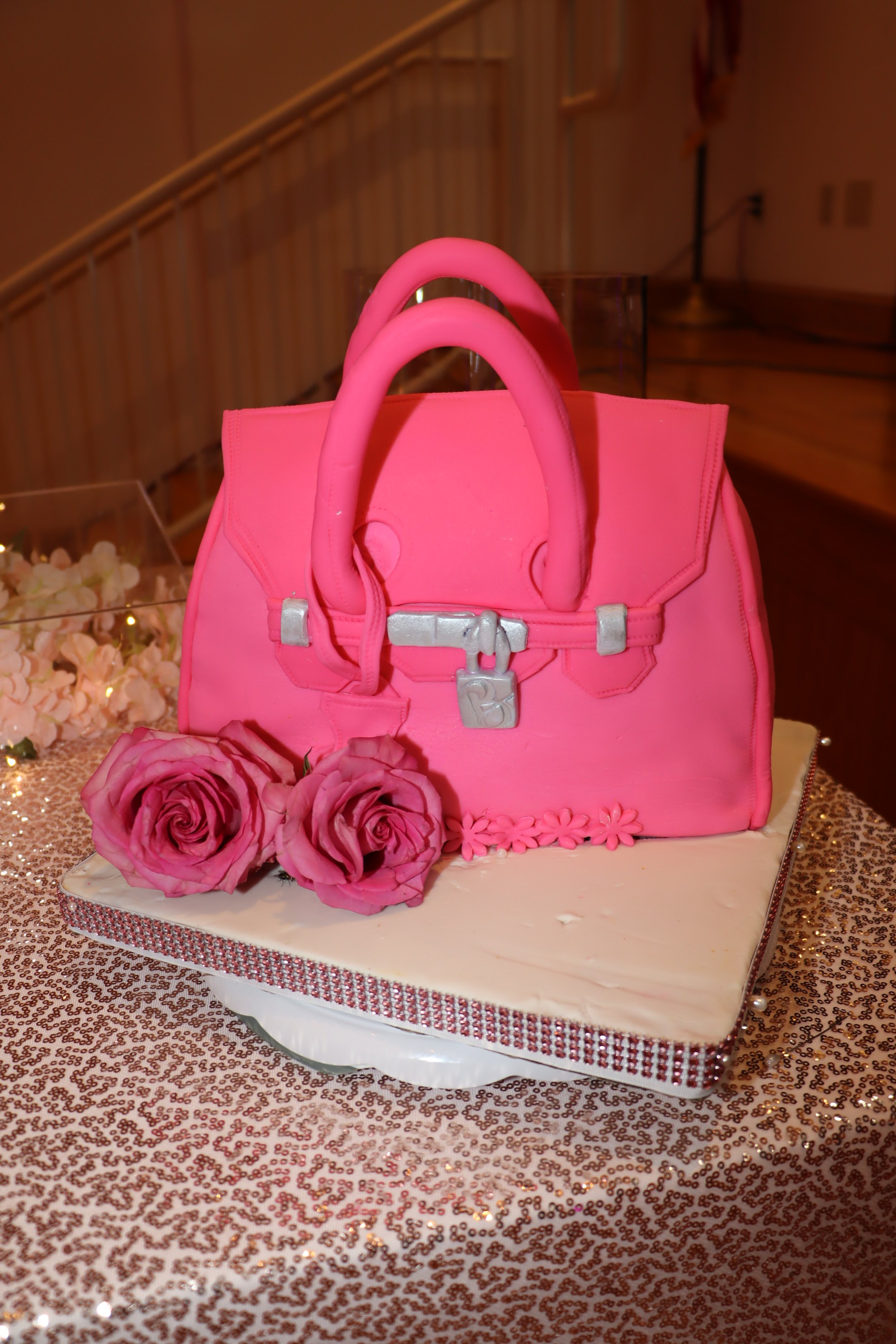 A cake designed to look like a pink handbag, decorated with roses and positioned on a glittery tablecloth.