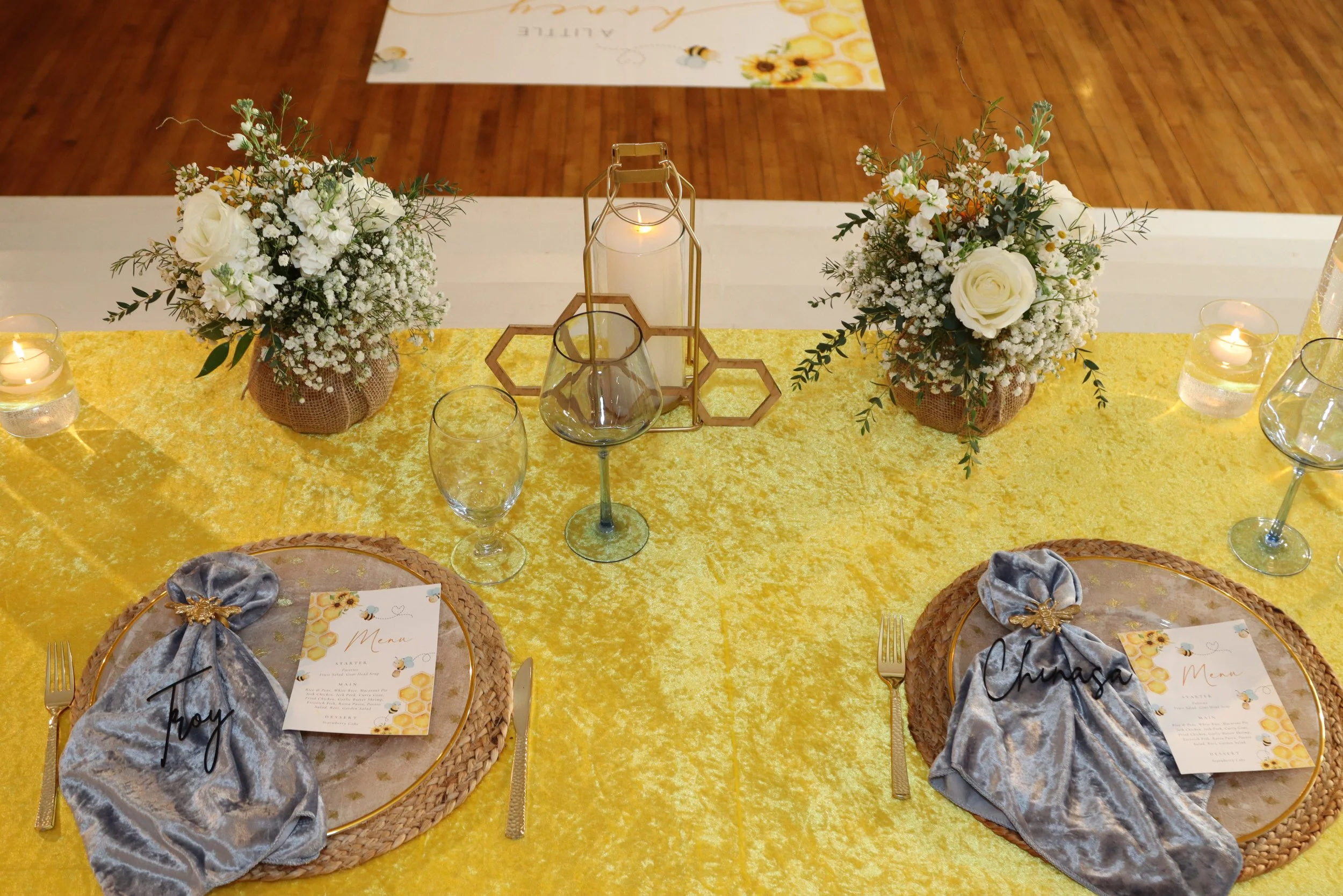 Decorated table for a special occasion with floral arrangements, candles, glasses, gold-rimmed plates, personalized napkins, and a menu card on each plate.
