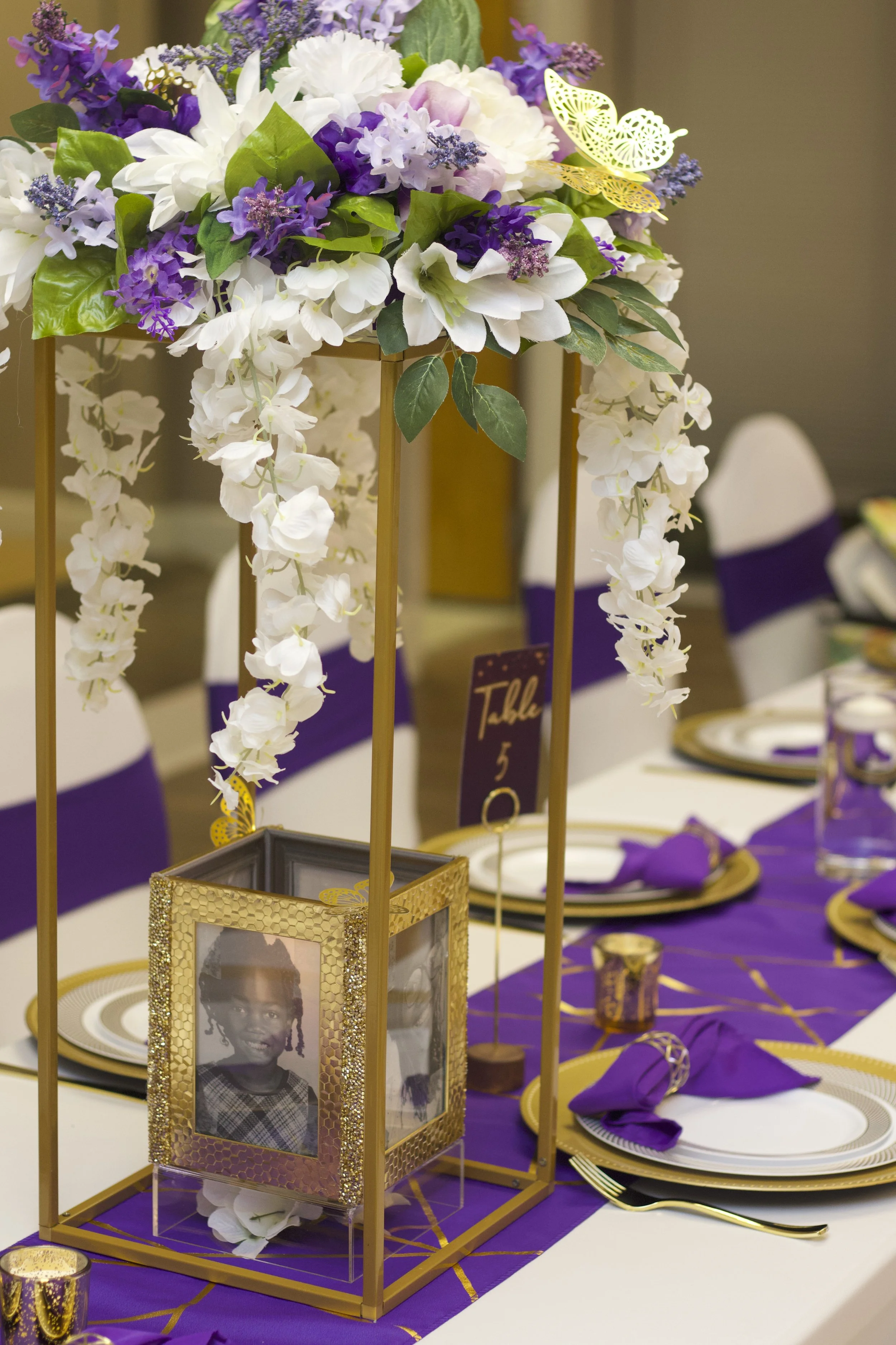 Elegant table centerpiece with purple and white flowers, greenery, and decorative butterflies, with framed photograph of a young girl, set for a formal event with purple and gold accents.