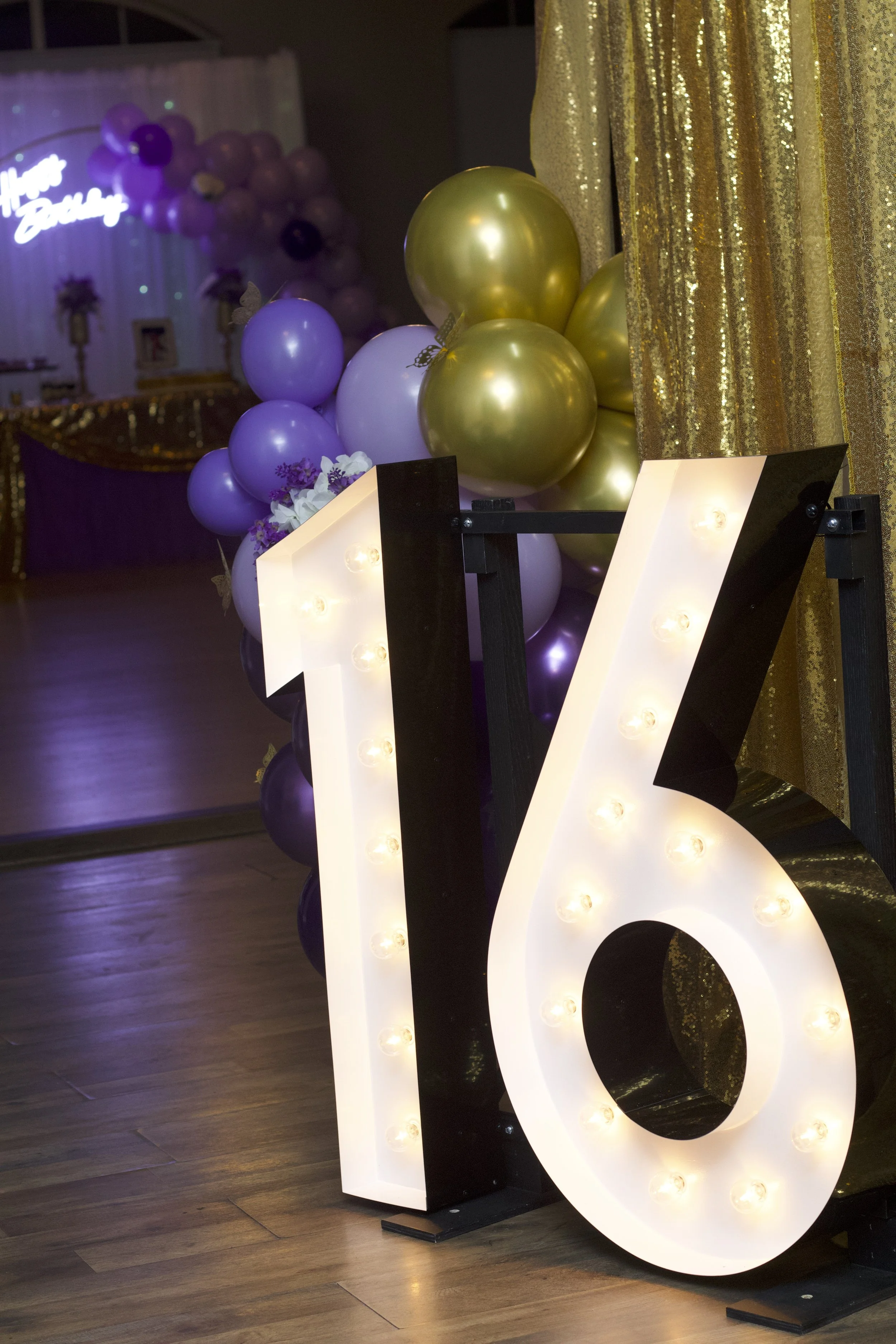 Decorative celebration setup with illuminated number 16 sign, purple, white, and gold balloons, gold curtain, and floral arrangements in the background.
