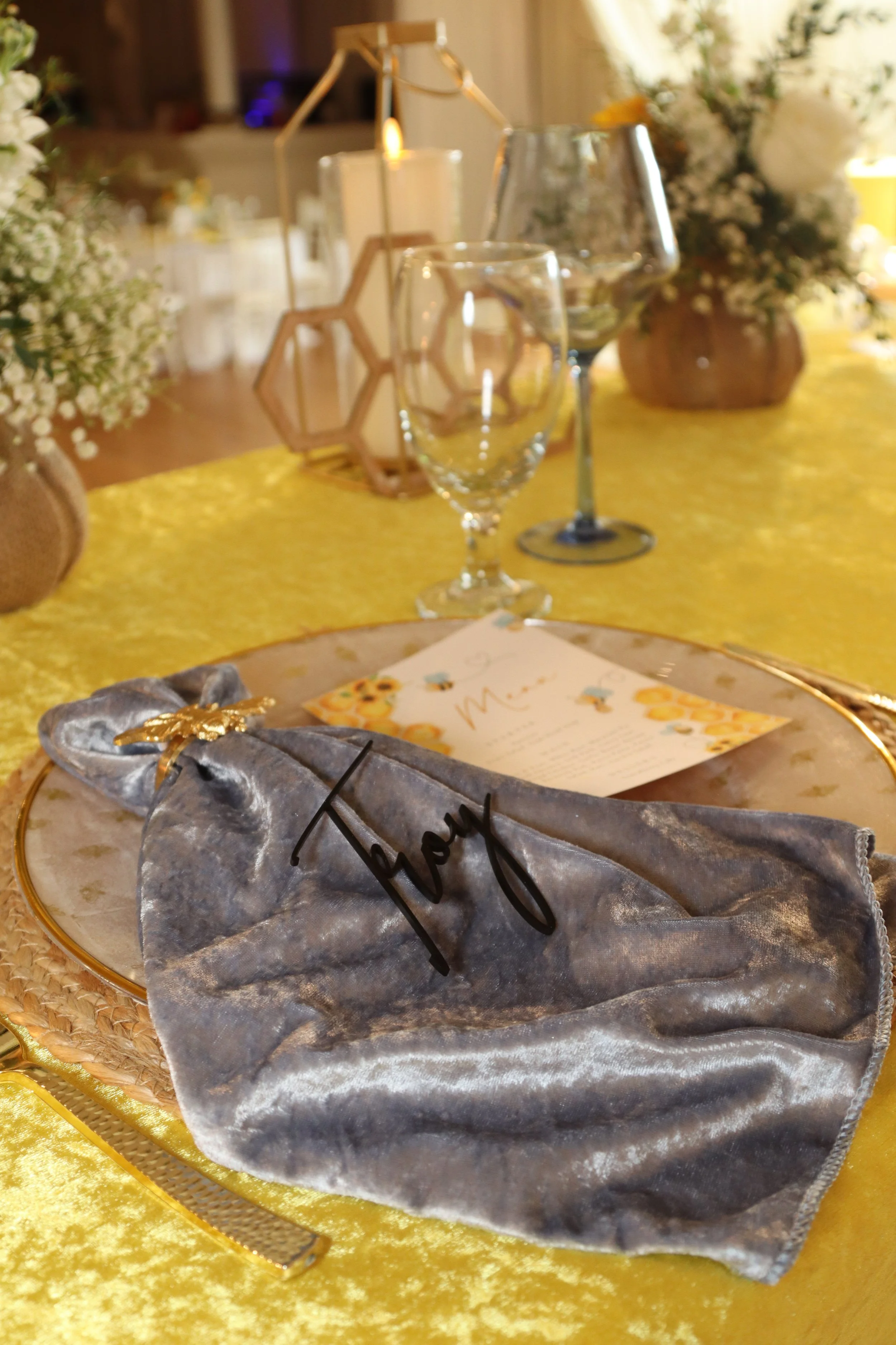 A silver napkin with the word 'Joy' written on it, placed on a decorative plate on a table with a yellow tablecloth, set for a celebration or dinner event. In the background were glasses, candles, and floral arrangements.