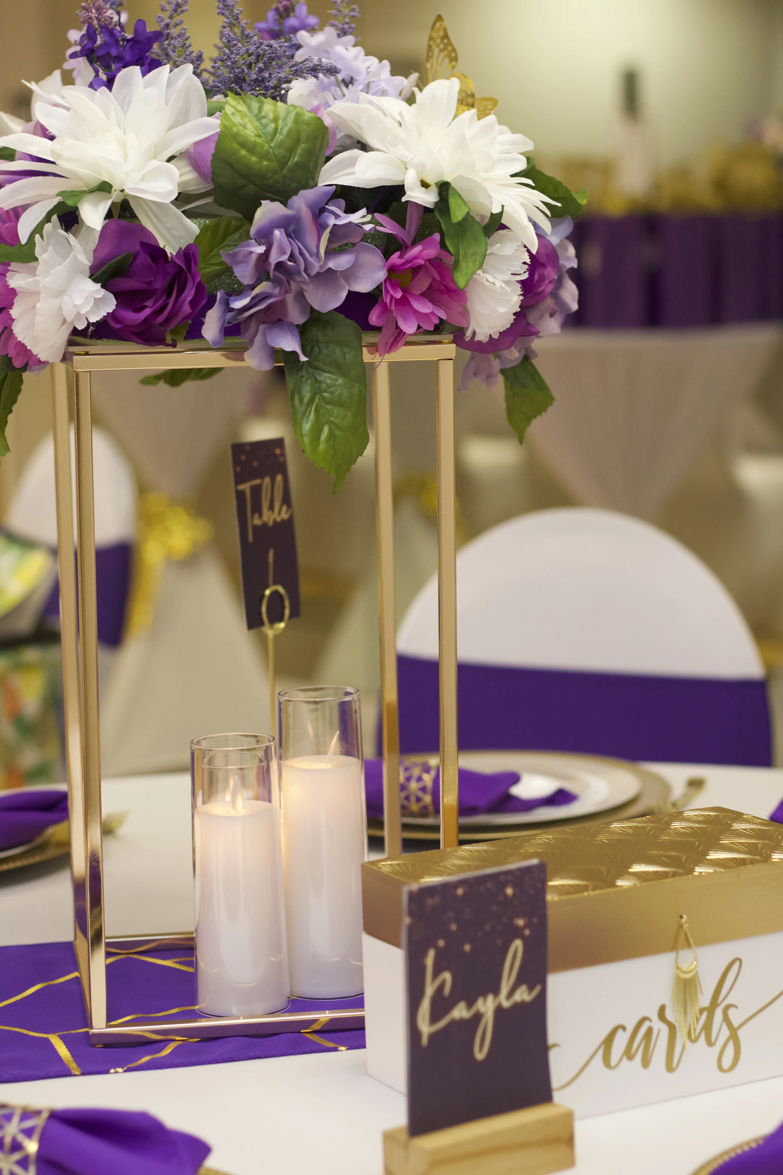 Decorated table with a floral centerpiece, candles in glass holders, purple and gold accents, and a table card with the name 'Kayla' in an elegant script.