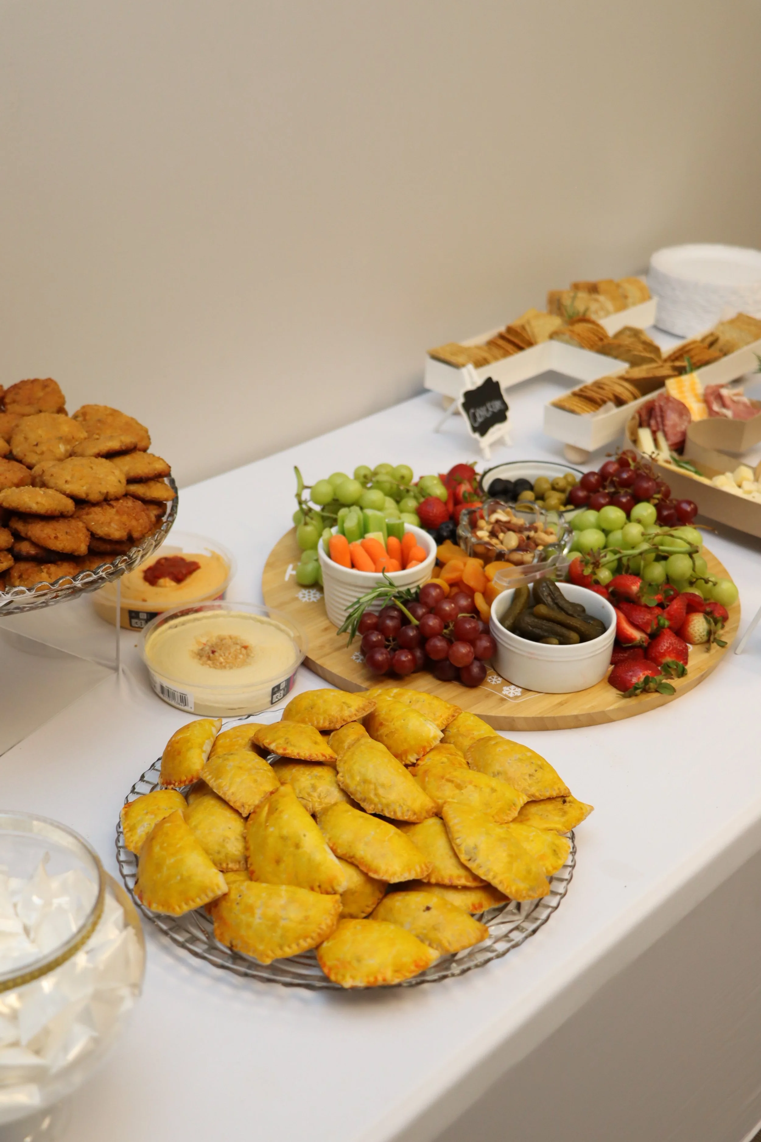 A variety of finger foods on a table, including a plate of crescent-shaped pastries, a platter of fresh grapes, strawberries, and pickles, bowls of dip, and trays of cookies and finger sandwiches.