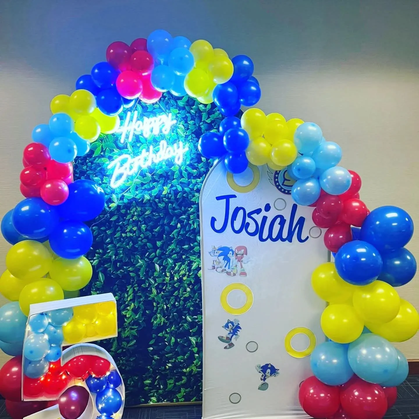 Colorful birthday decoration with balloons in red, yellow, blue, and light blue, surrounding a backdrop with a Neon 'Happy Birthday' sign and a 'Sonic the Hedgehog'-themed birthday banner with the name Josiah.