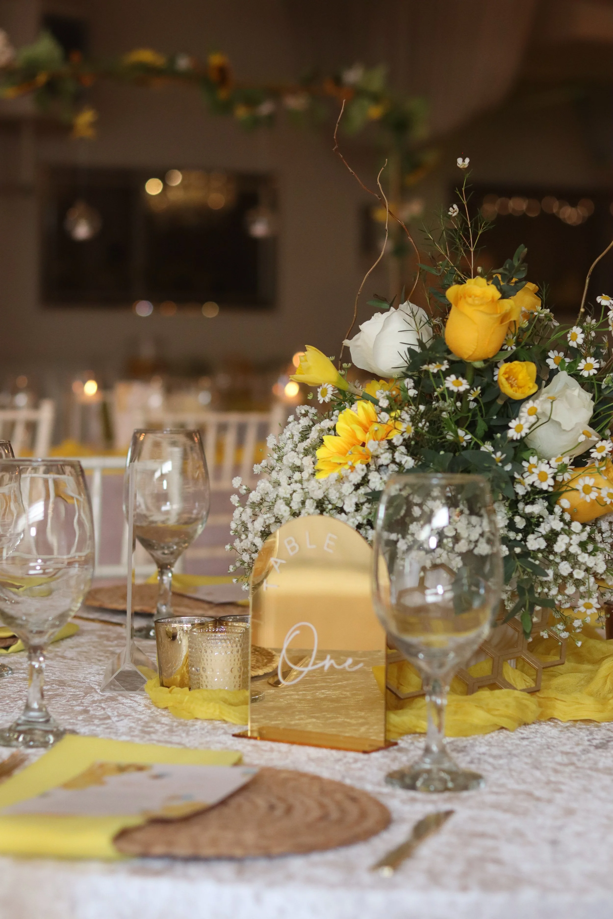 Elegant table setting with wine glasses, a floral centerpiece featuring yellow and white flowers, a mirror with the words "TABLE One," and yellow accents, set for a celebration or wedding reception.